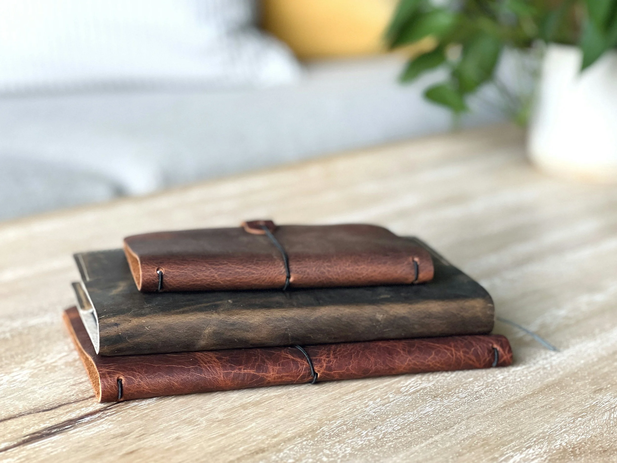 Stack of three leather-bound notebooks or journals on a wooden surface, with a blurry background including a white vase and green plant leaves.