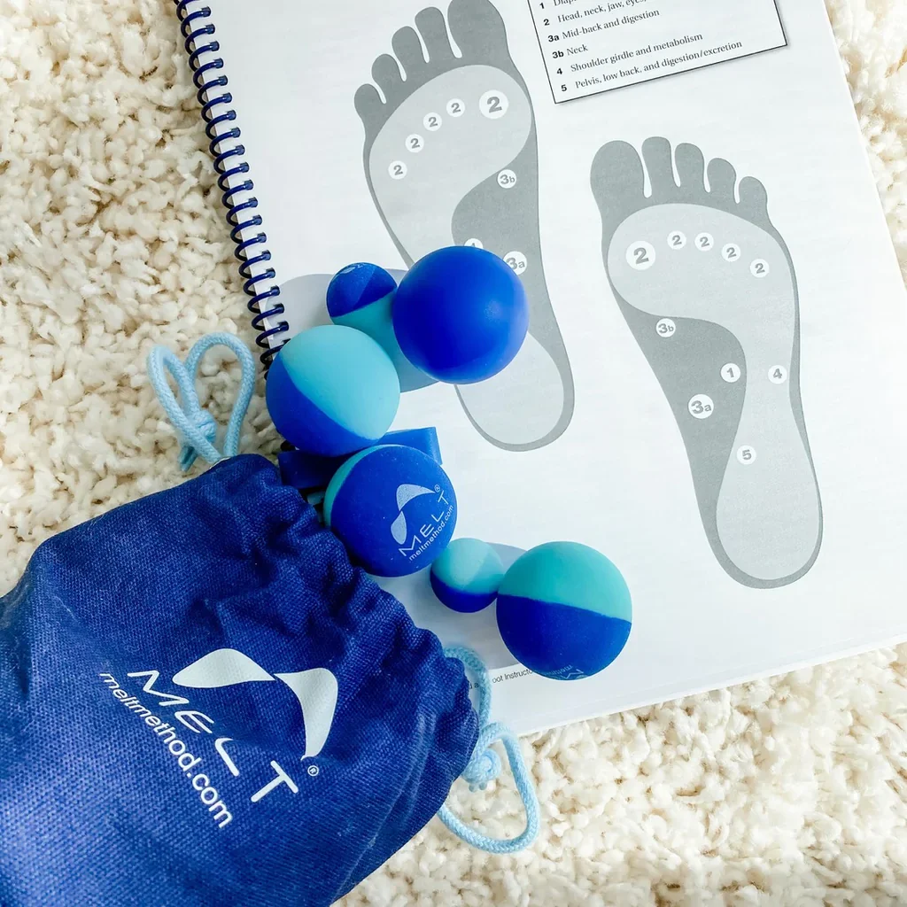 Foam roller balls in various shades of blue on a white carpet, with a blue drawstring bag labeled 'MEET' and a laminated foot reflexology chart.