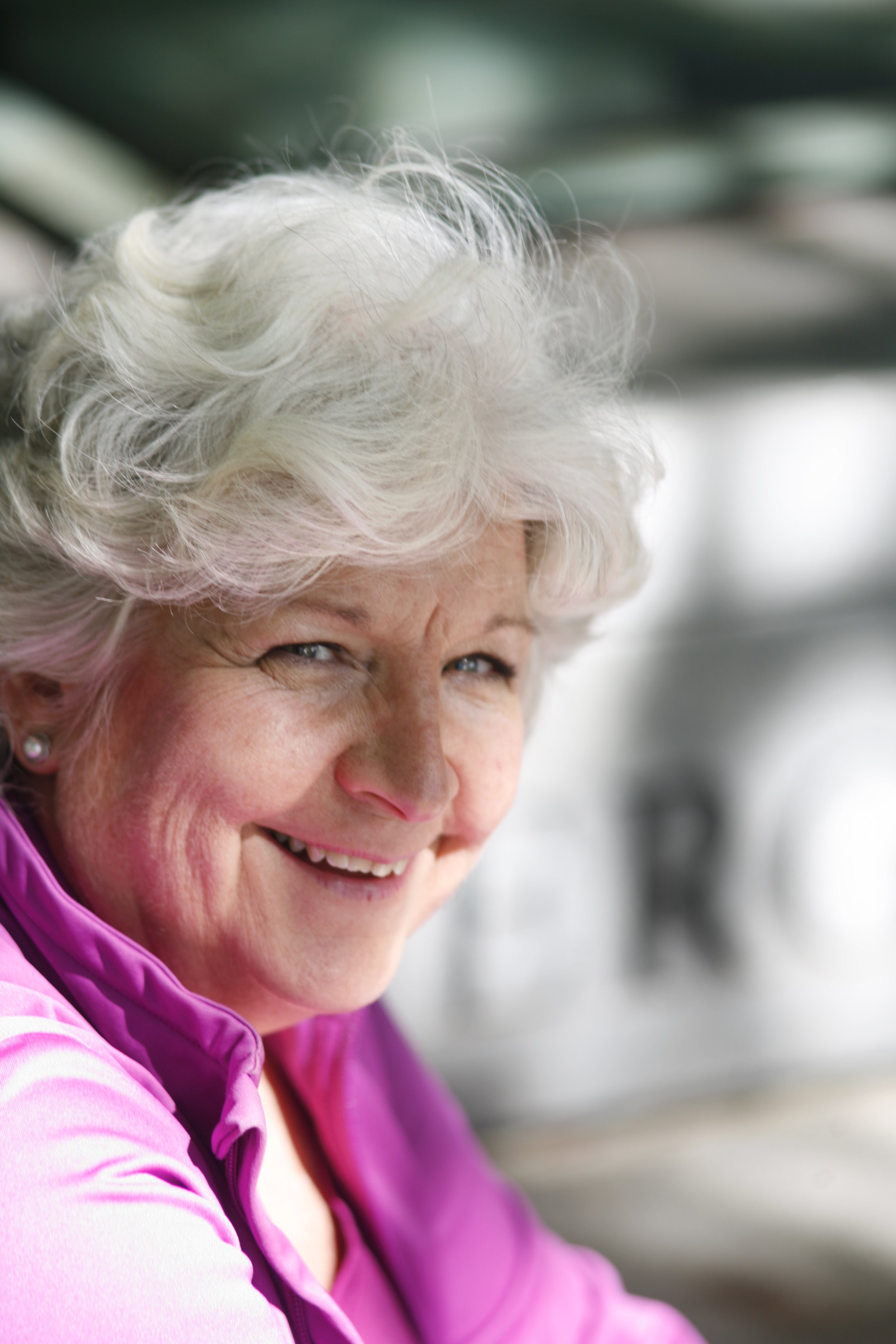 A smiling elderly woman with curly white hair, wearing a purple jacket.