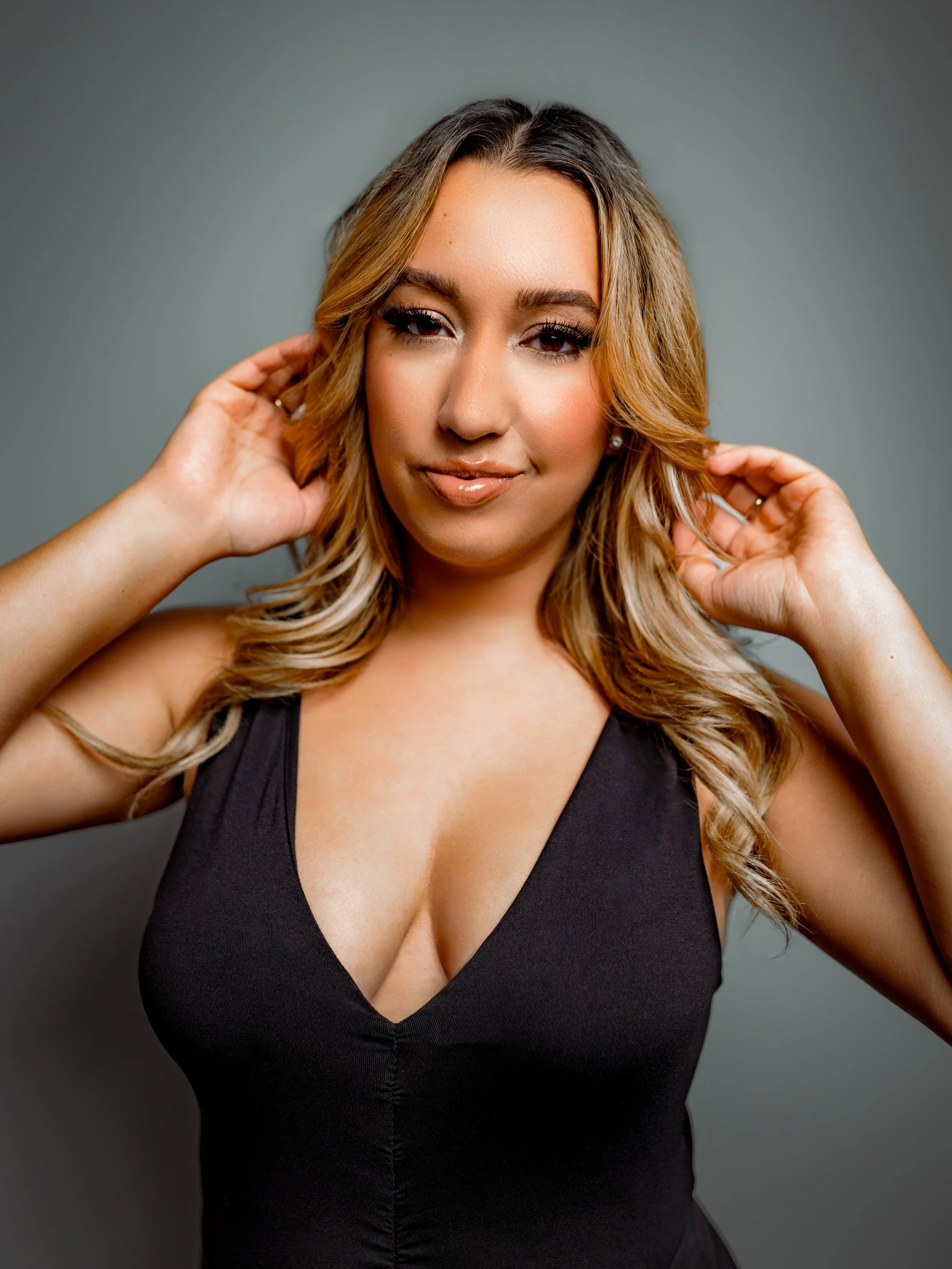 A portrait of a woman with blonde and brown hair, wearing a black sleeveless top with a deep neckline, posing against a gray background.