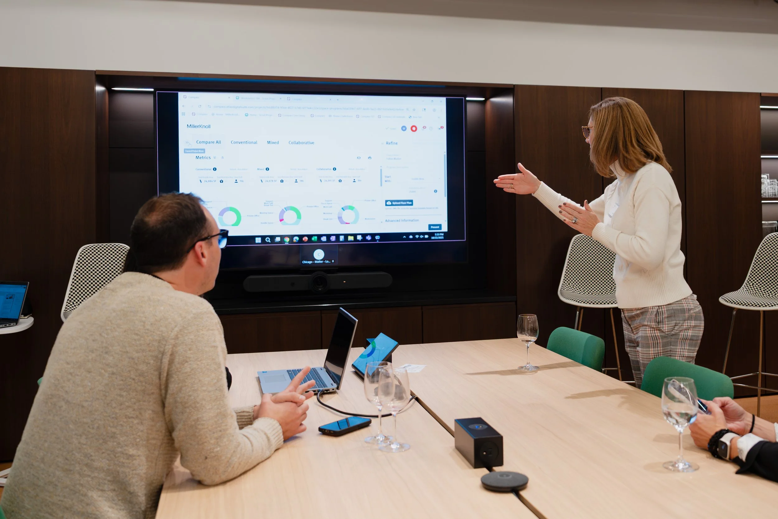 A woman presenting a chart on a large screen to three seated people in a conference room.