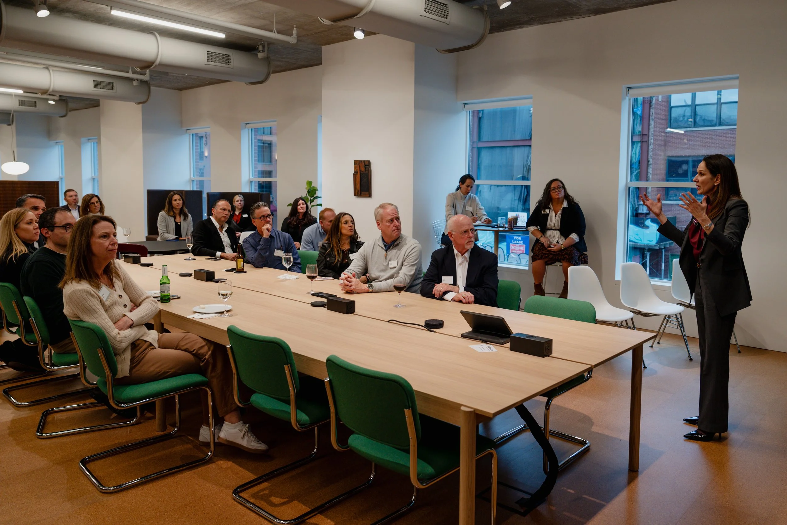 A woman in a business suit is speaking to a group of people seated around a long conference table in a modern meeting room. The audience appears engaged, with some taking notes and others listening attentively.