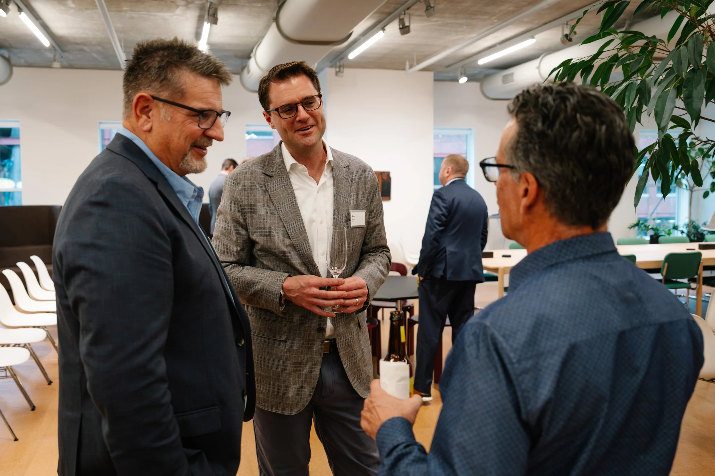Three men in business attire having a conversation at a networking event, with drinks in their hands in a modern office space.