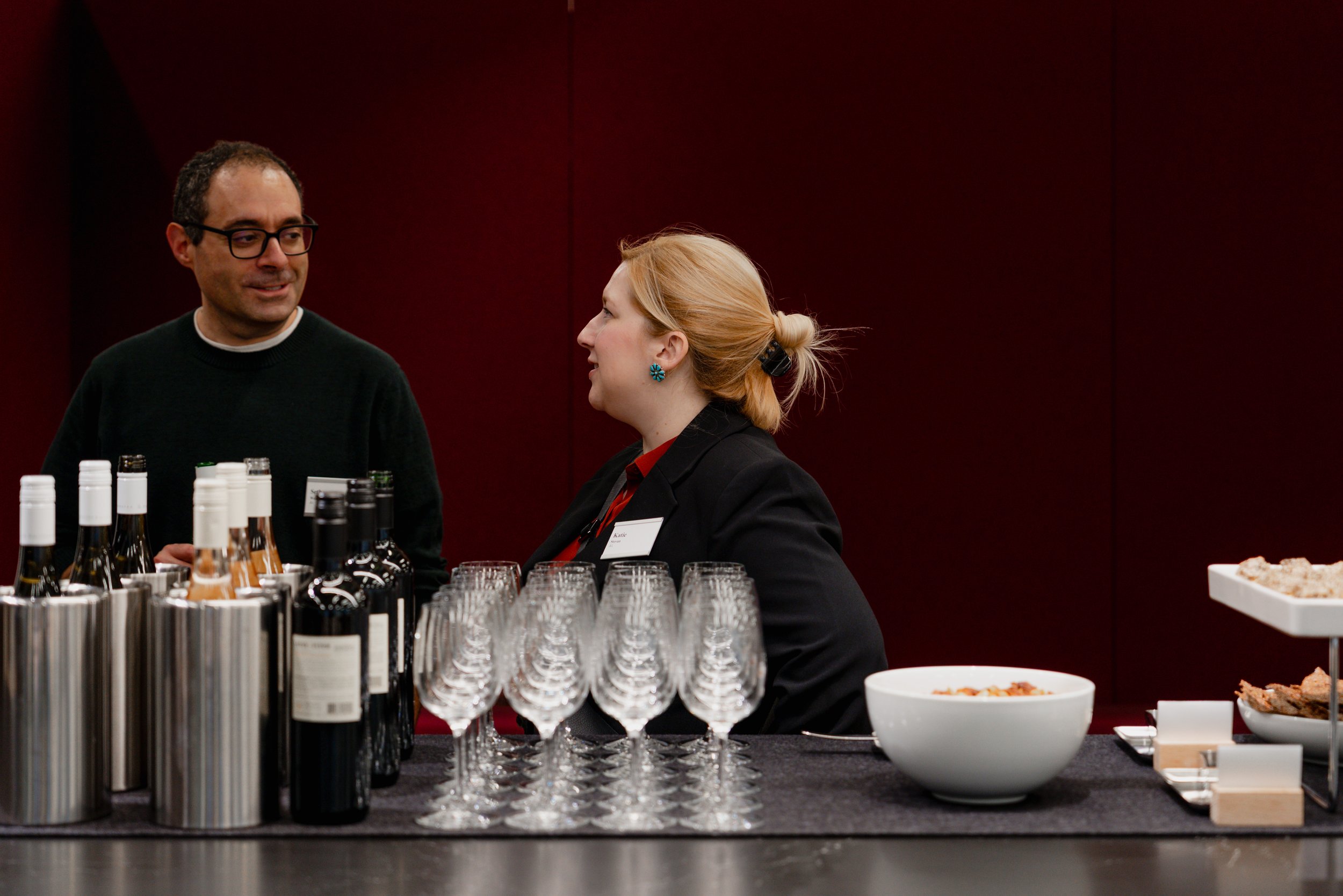 A man and woman having a conversation at a bar with wine bottles, glasses, and snacks on the counter.