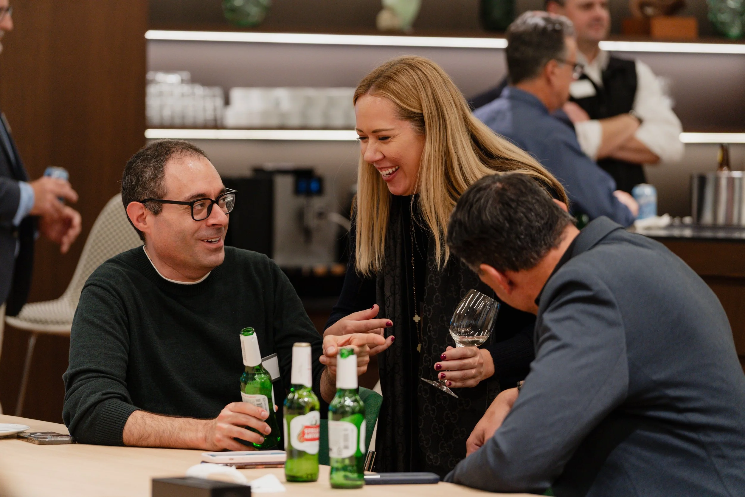 People at a social gathering enjoying drinks and conversation, with three men seated at a table and a woman standing and smiling, holding a wine glass.