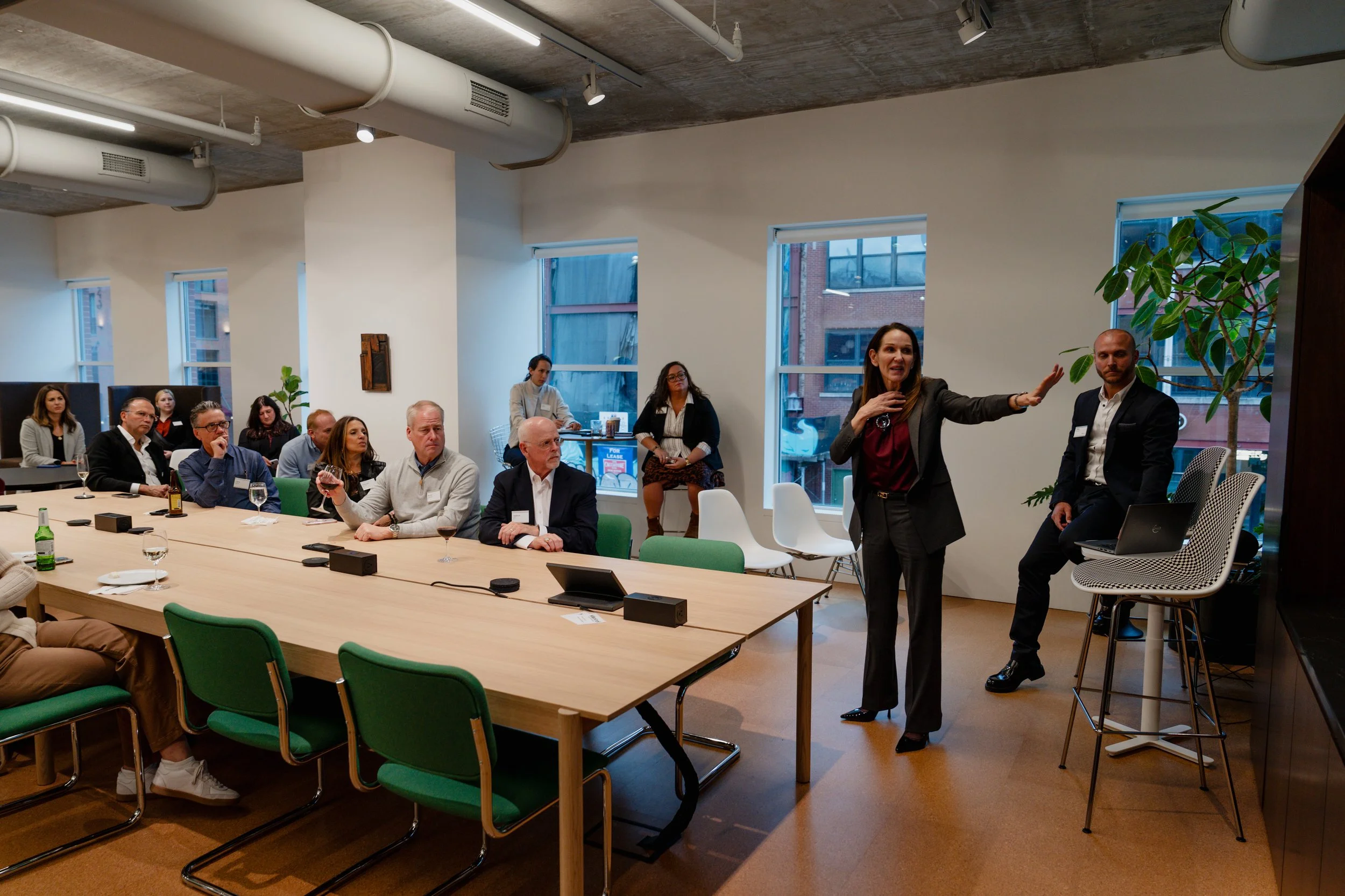A woman giving a presentation to a group of business professionals in a modern conference room with floor-to-ceiling windows, potted plants, and a large wooden table.