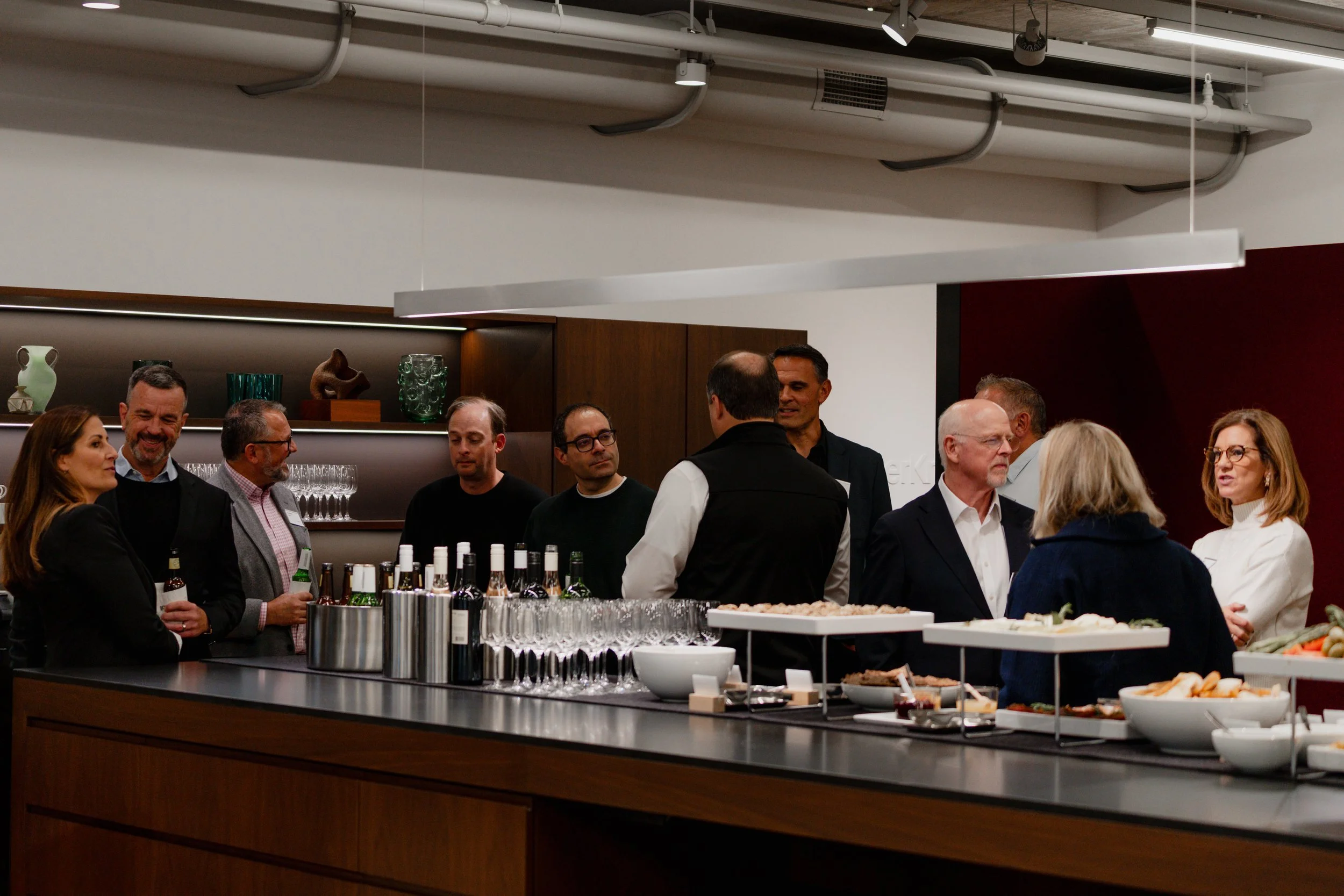 Group of people gathered in a modern kitchen or event space, engaging in conversation near a table with food and drinks, including wine bottles, glasses, and appetizers.
