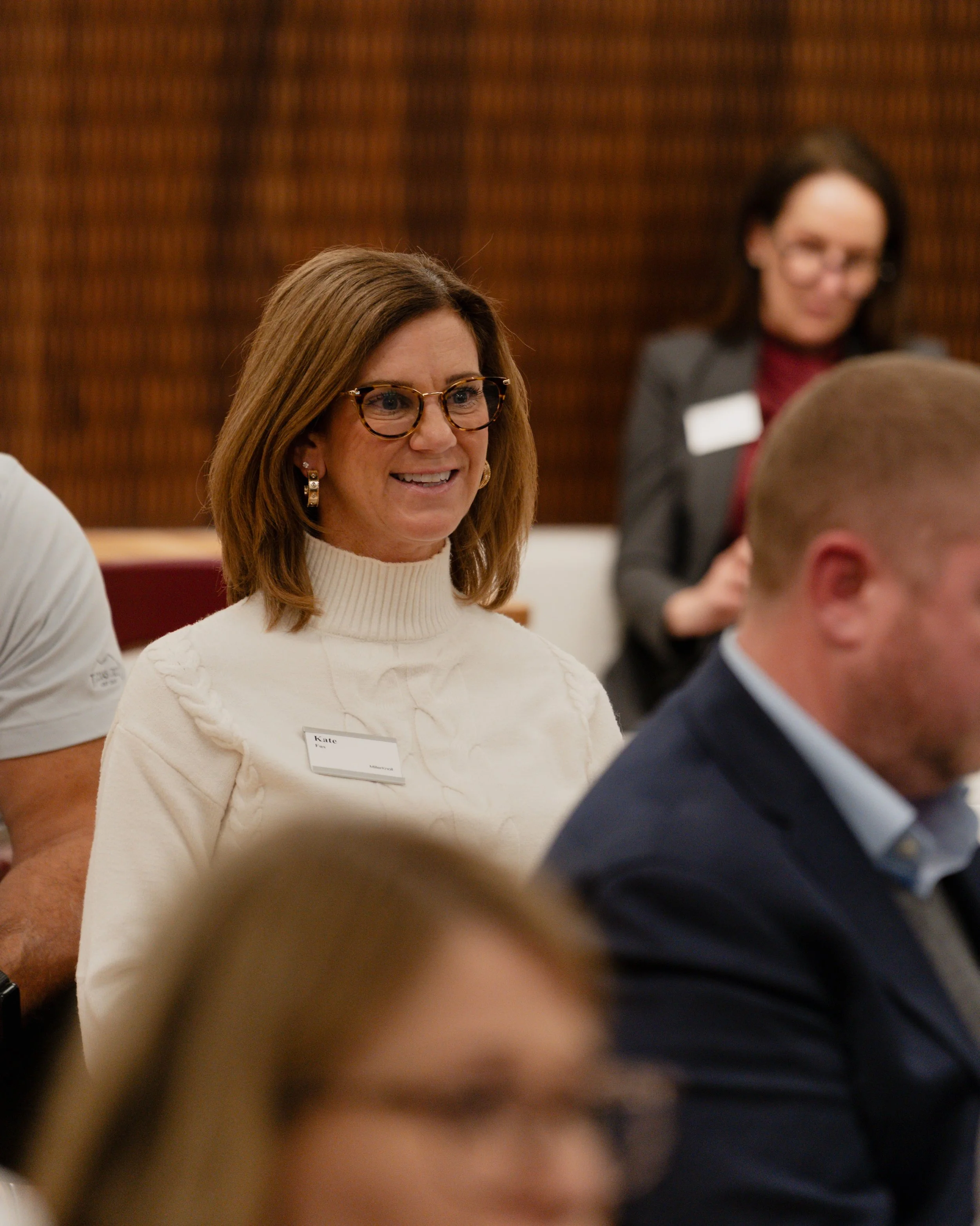A woman with shoulder-length brown hair, glasses, wearing a white turtleneck sweater and a name tag, smiling at a professional event.