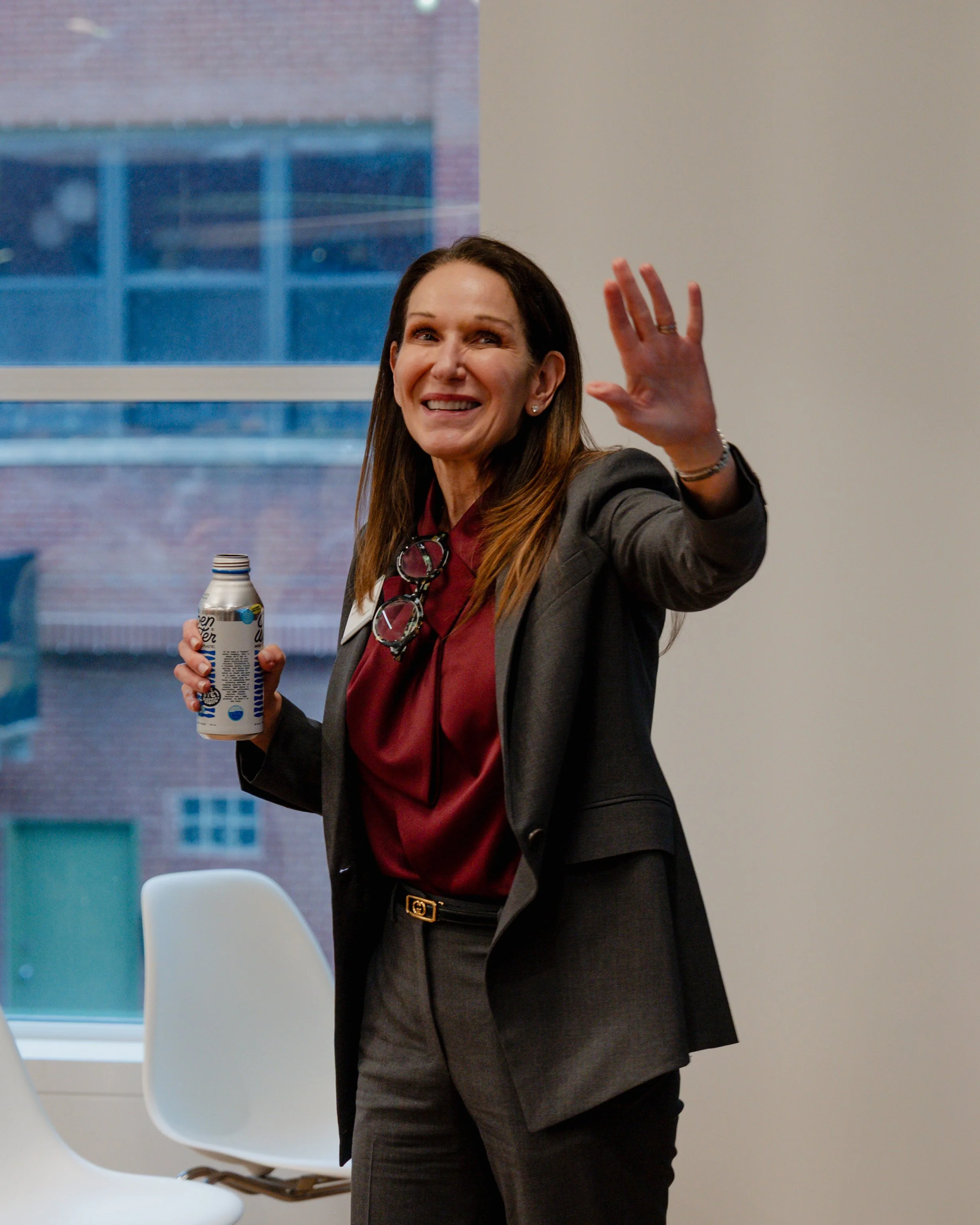 A woman with long brown hair smiling and waving, holding a can of soda, dressed in a gray blazer and burgundy blouse, standing indoors near a window.