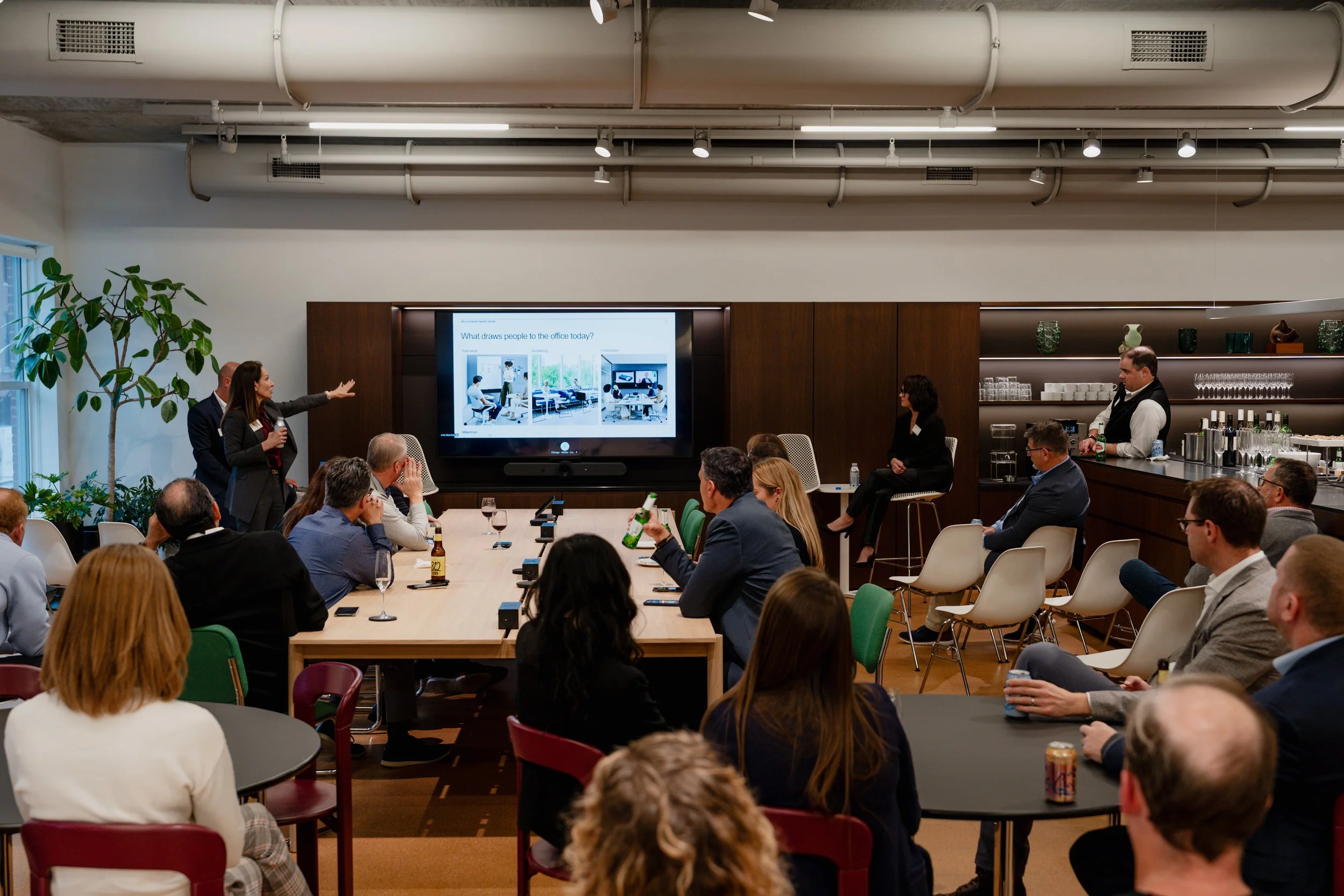 Business presentation in a conference room with a woman speaker gesturing towards a large screen displaying a slide about office activity. Audience members are seated at tables, some holding drinks, and a bartender is working behind a bar on the righ
