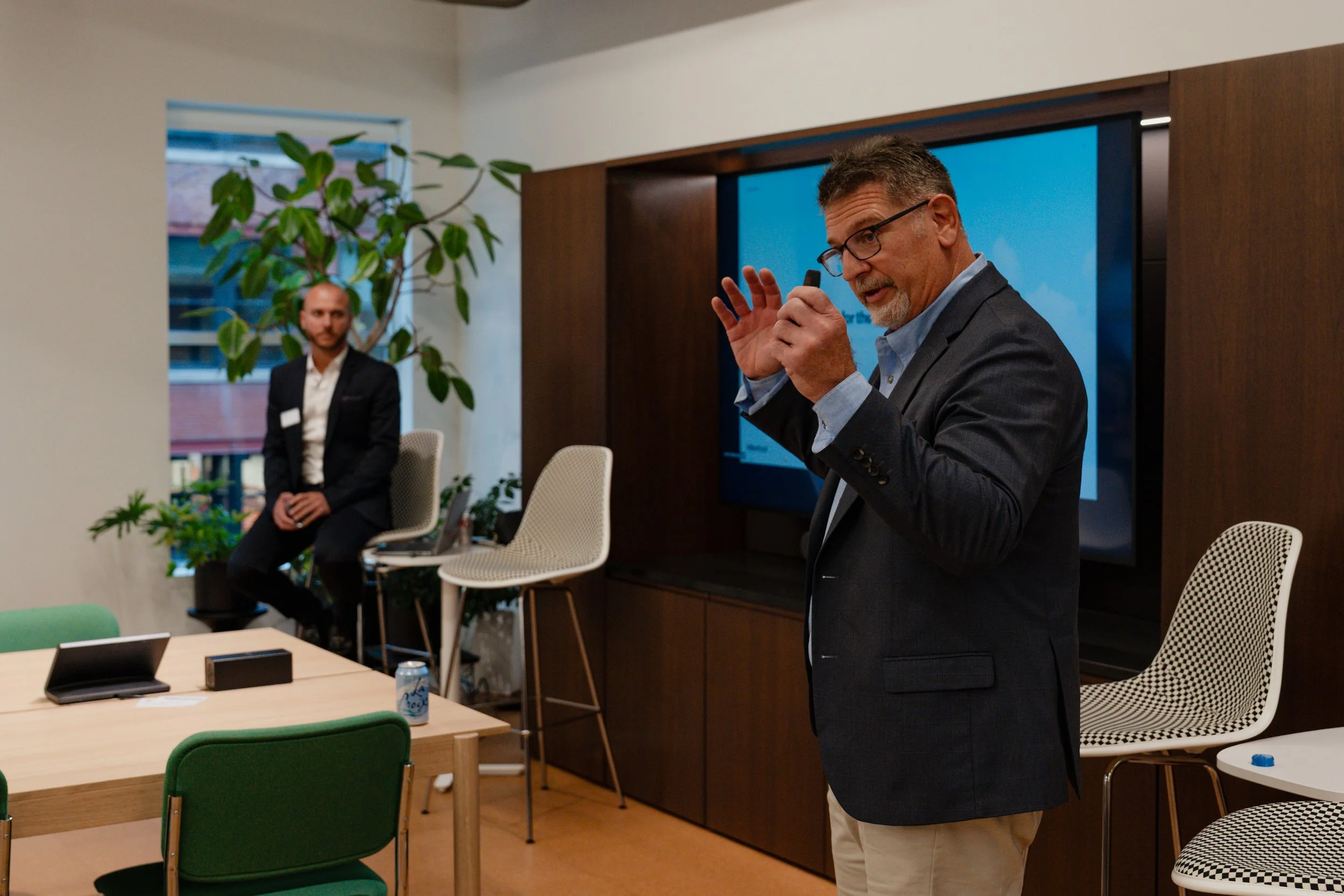 A man with gray hair, beard, and glasses giving a presentation in a conference room, with another man sitting on a high chair in the background near a window and a large TV screen on the wall.