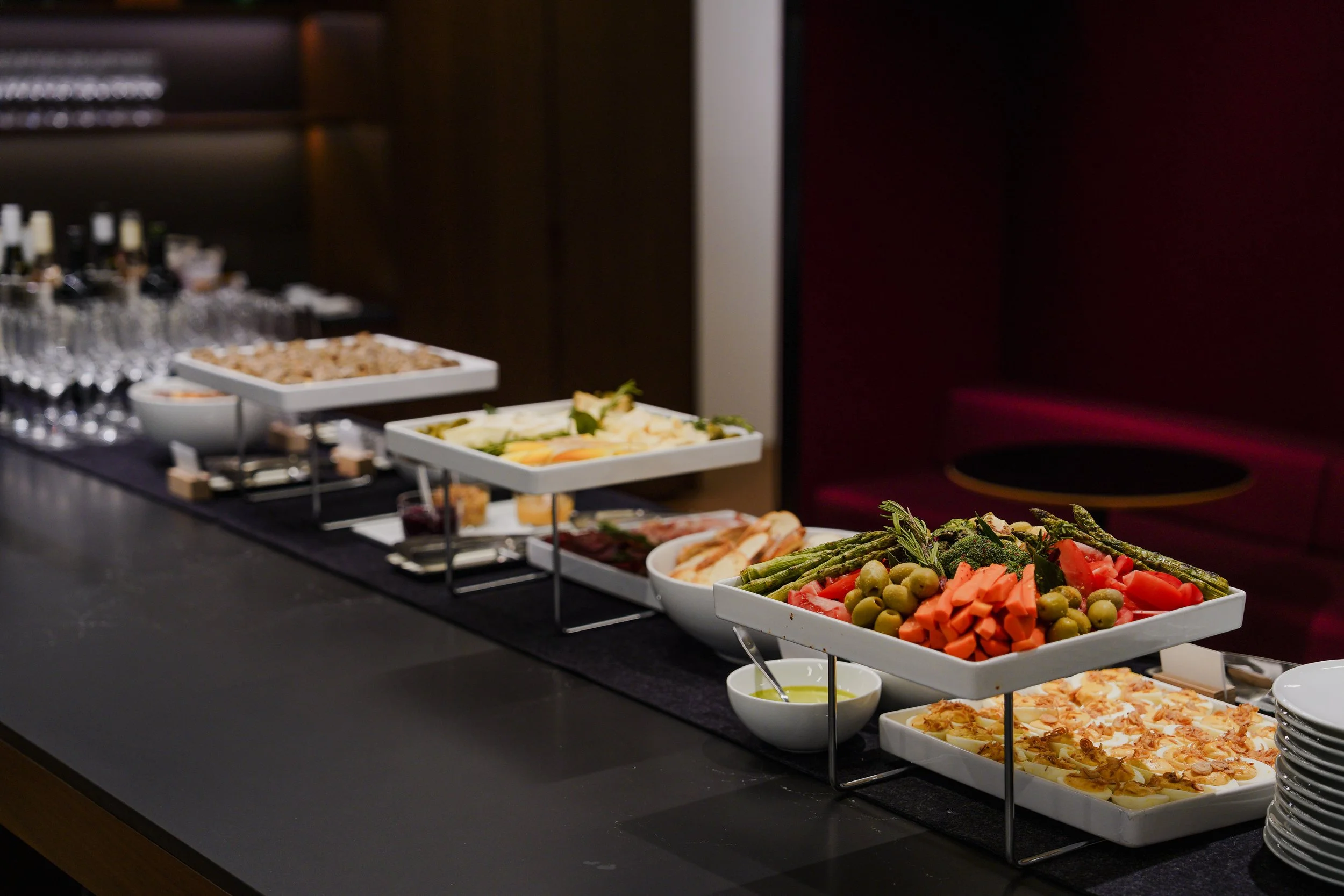 Buffet table with a variety of salads, vegetables, and appetizers, including sliced meats, olives, cherry tomatoes, carrots, asparagus, and broccoli, with wine glasses and bottles in the background.