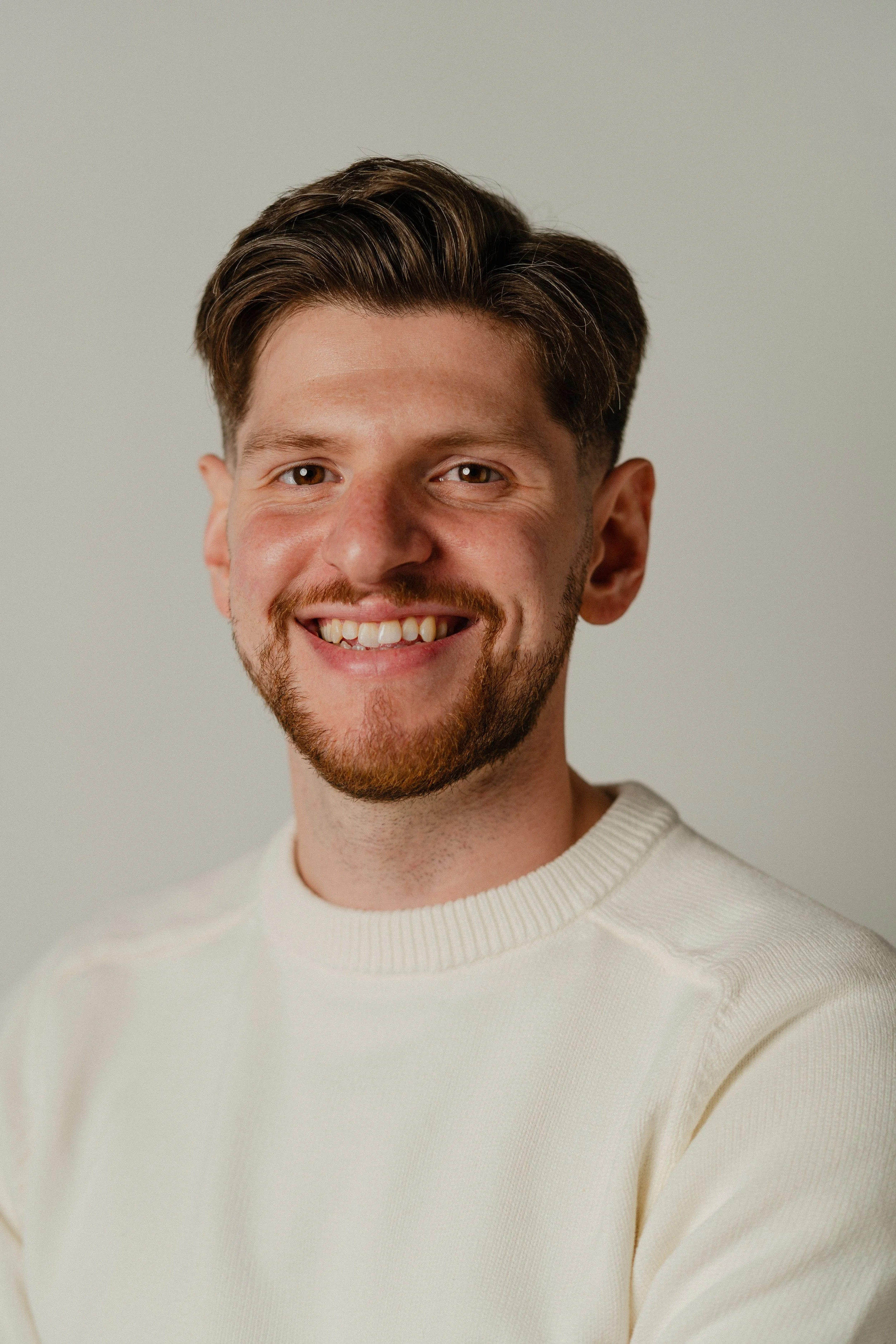 A portrait of a young man with brown hair and a beard, smiling and wearing a cream-colored sweater, against a neutral background.