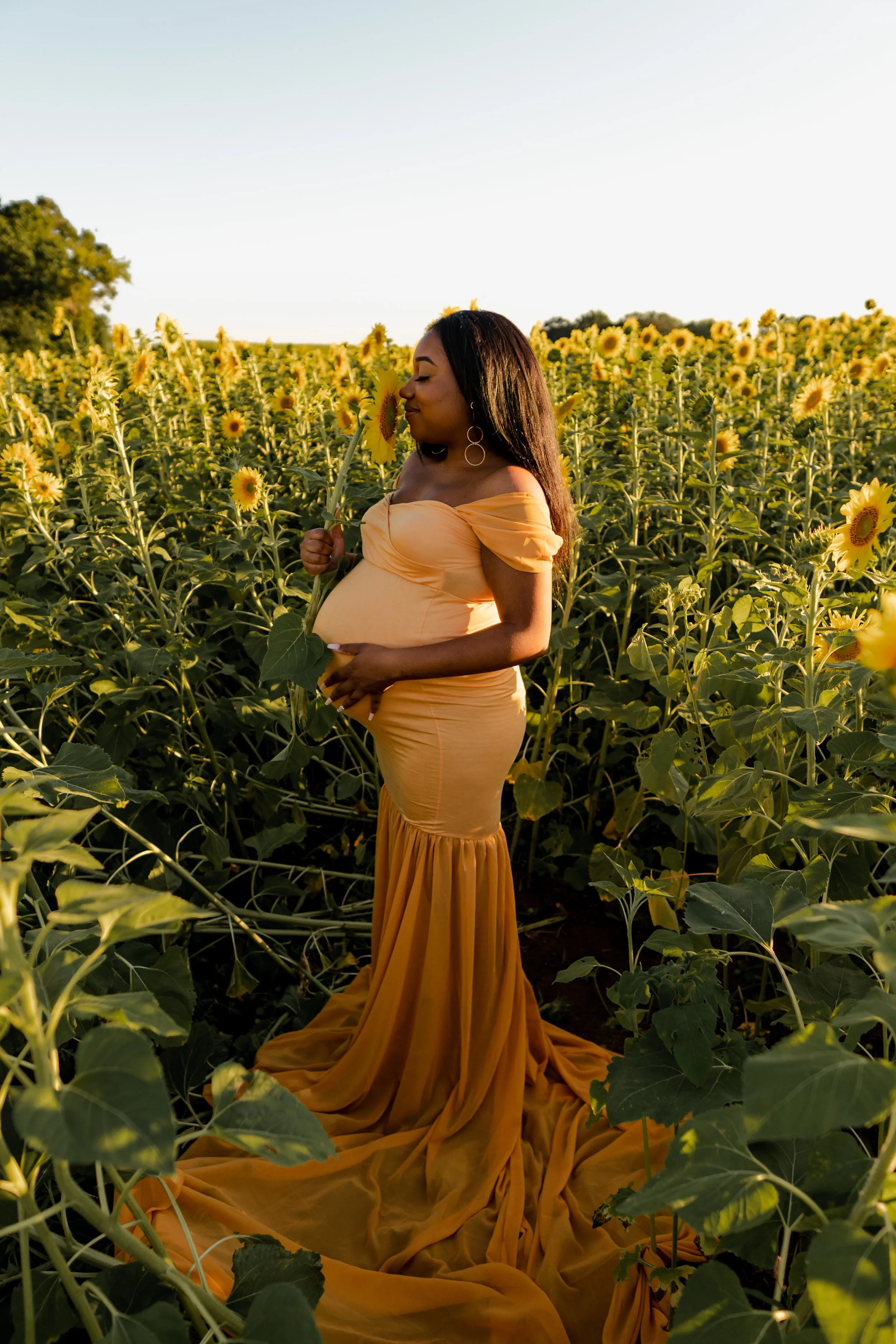 A portrait of a pregnant woman in a yellow off-shoulder gown standing in a sunflower field, holding a sunflower close to her face, with her eyes closed.