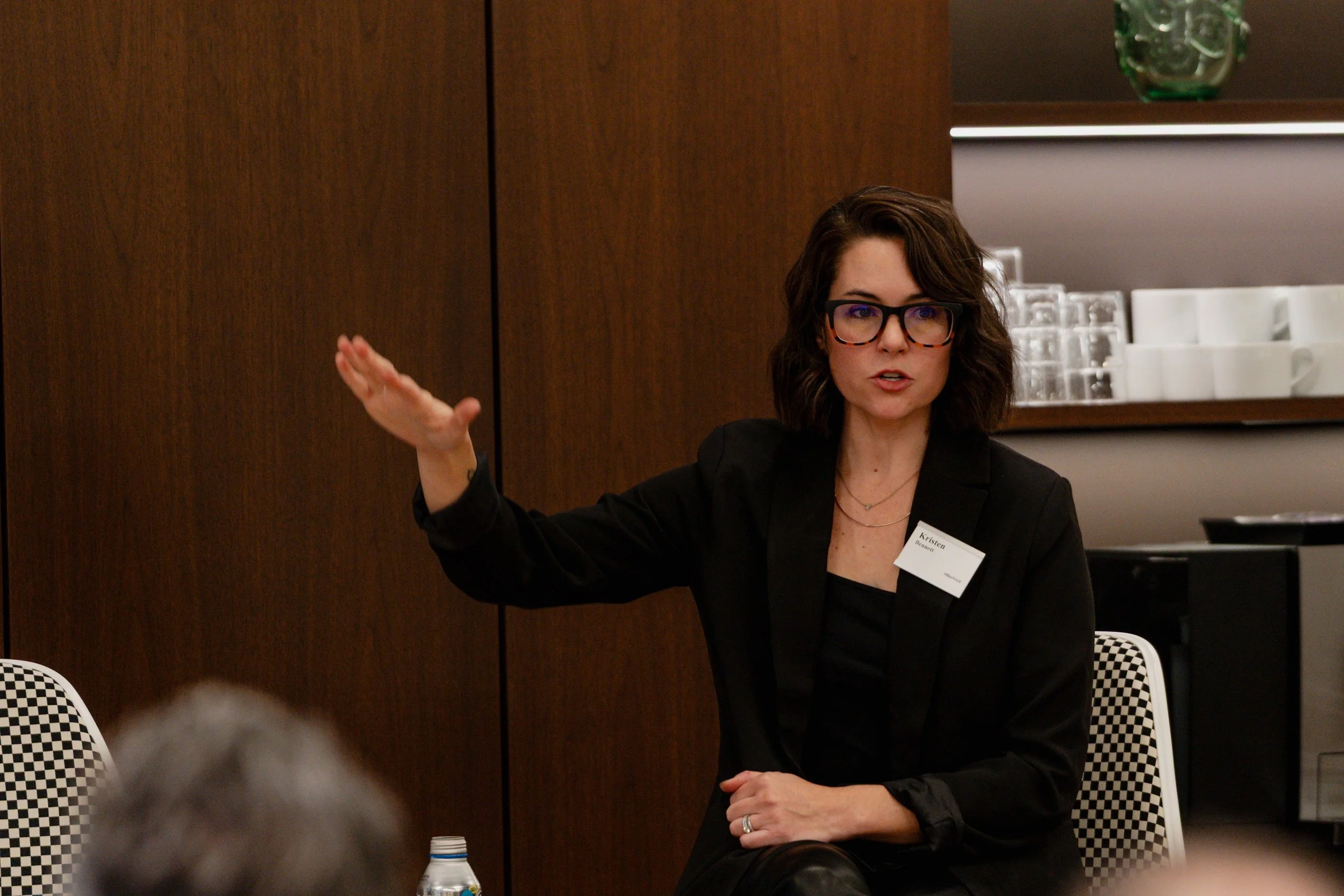 A woman with shoulder-length curly brown hair, wearing glasses and a black blazer, is speaking and gesturing with her right hand during a discussion or presentation in a conference room.