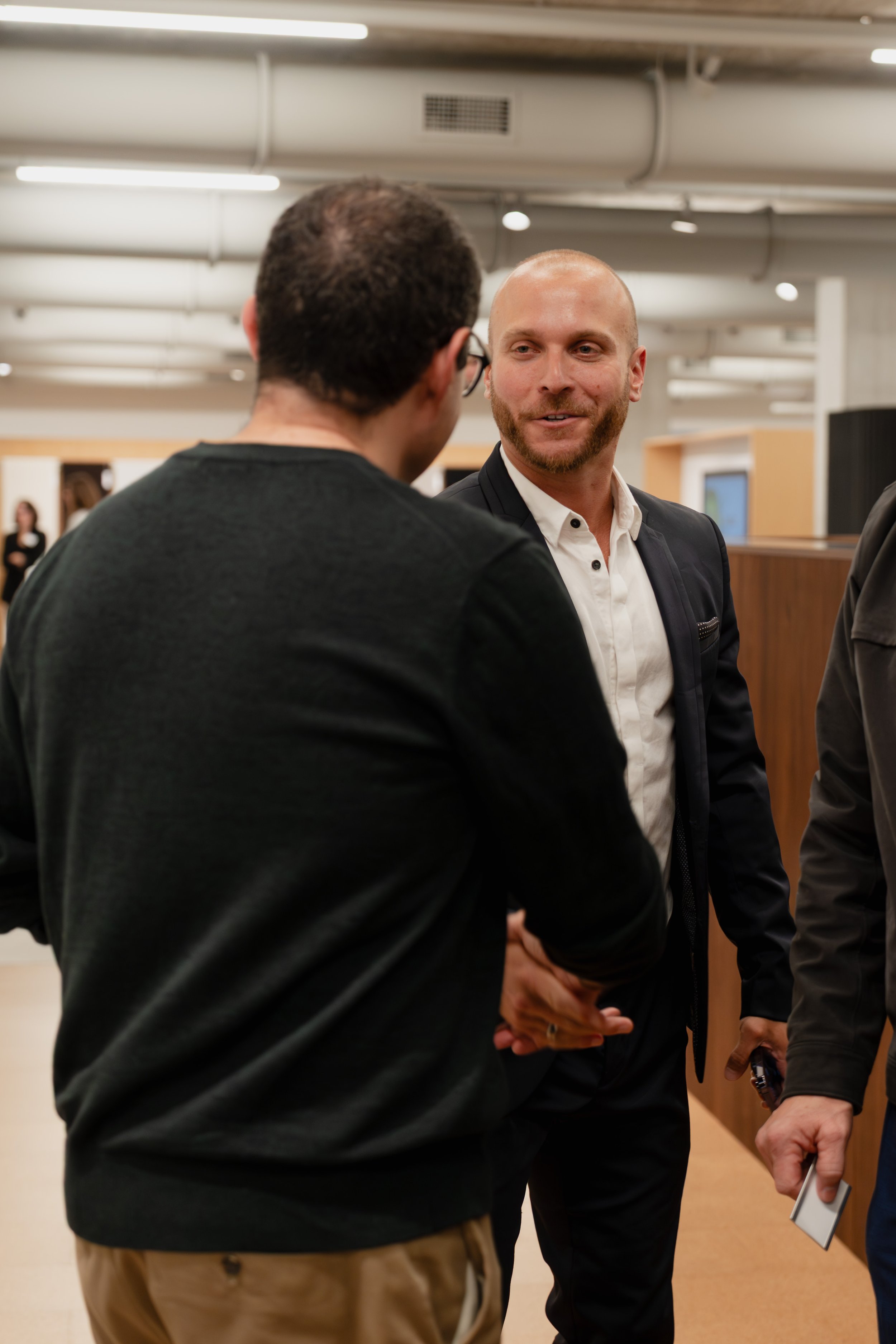Two men are engaged in a conversation, shaking hands in an indoor setting, with other people and a wooden divider in the background.