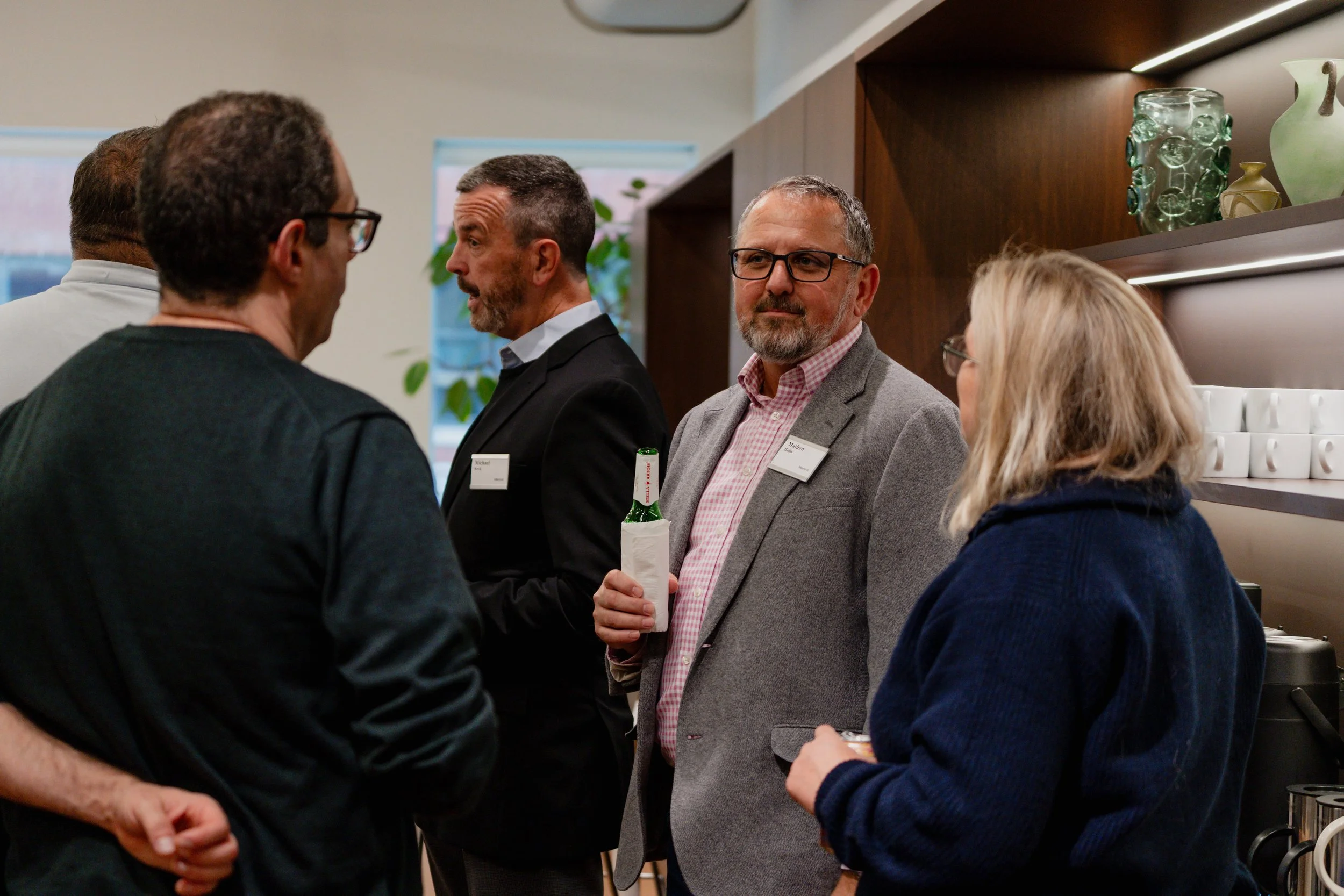 Group of five adults engaged in conversation at a networking event or conference, standing indoors near a wooden shelf with decorative glassware and coffee cups.