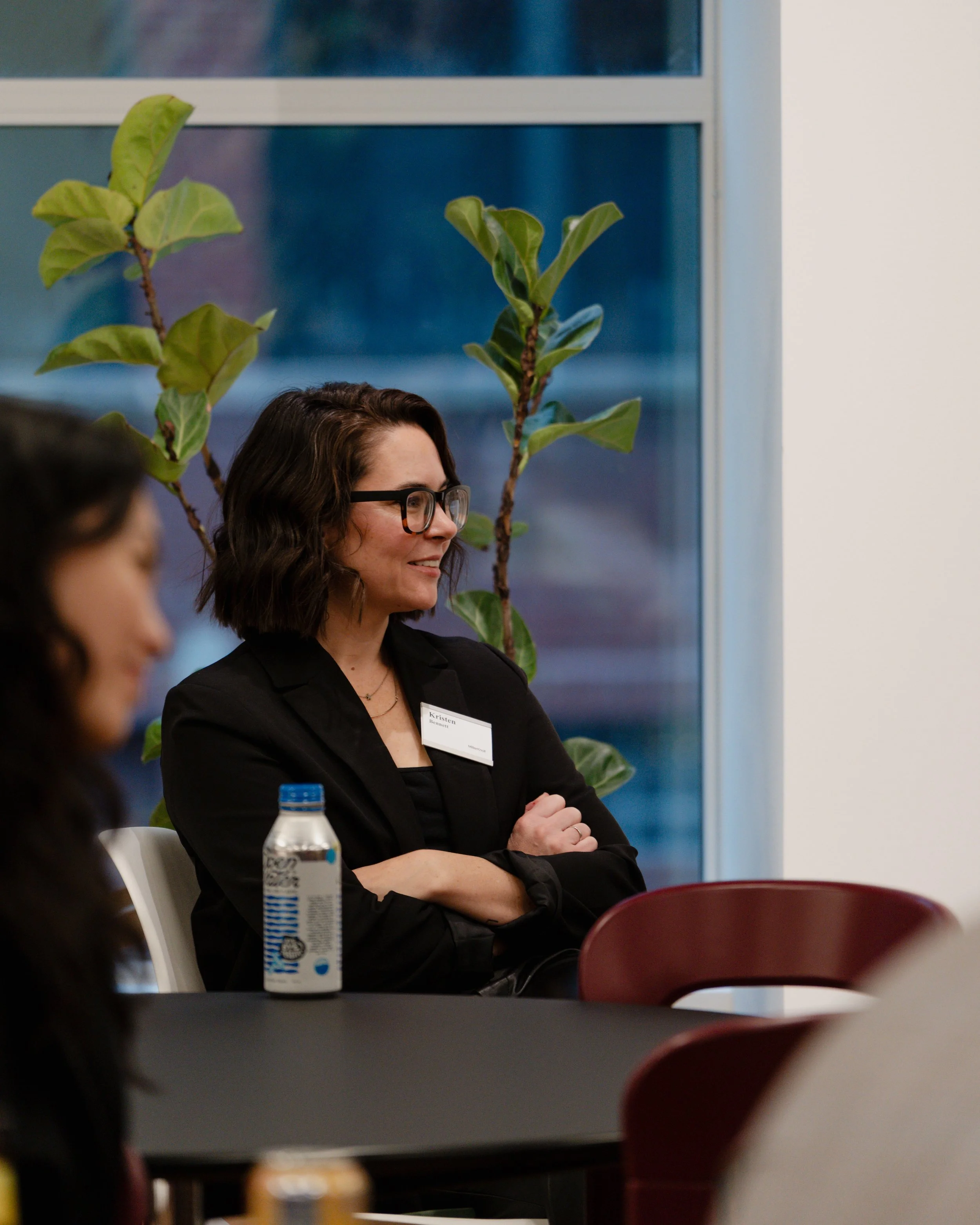 A woman with dark curly hair, glasses, and a black blazer is sitting at a table. She is smiling and has a name tag. There is a water bottle in front of her. Large green plants are visible behind her, and a window is in the background.
