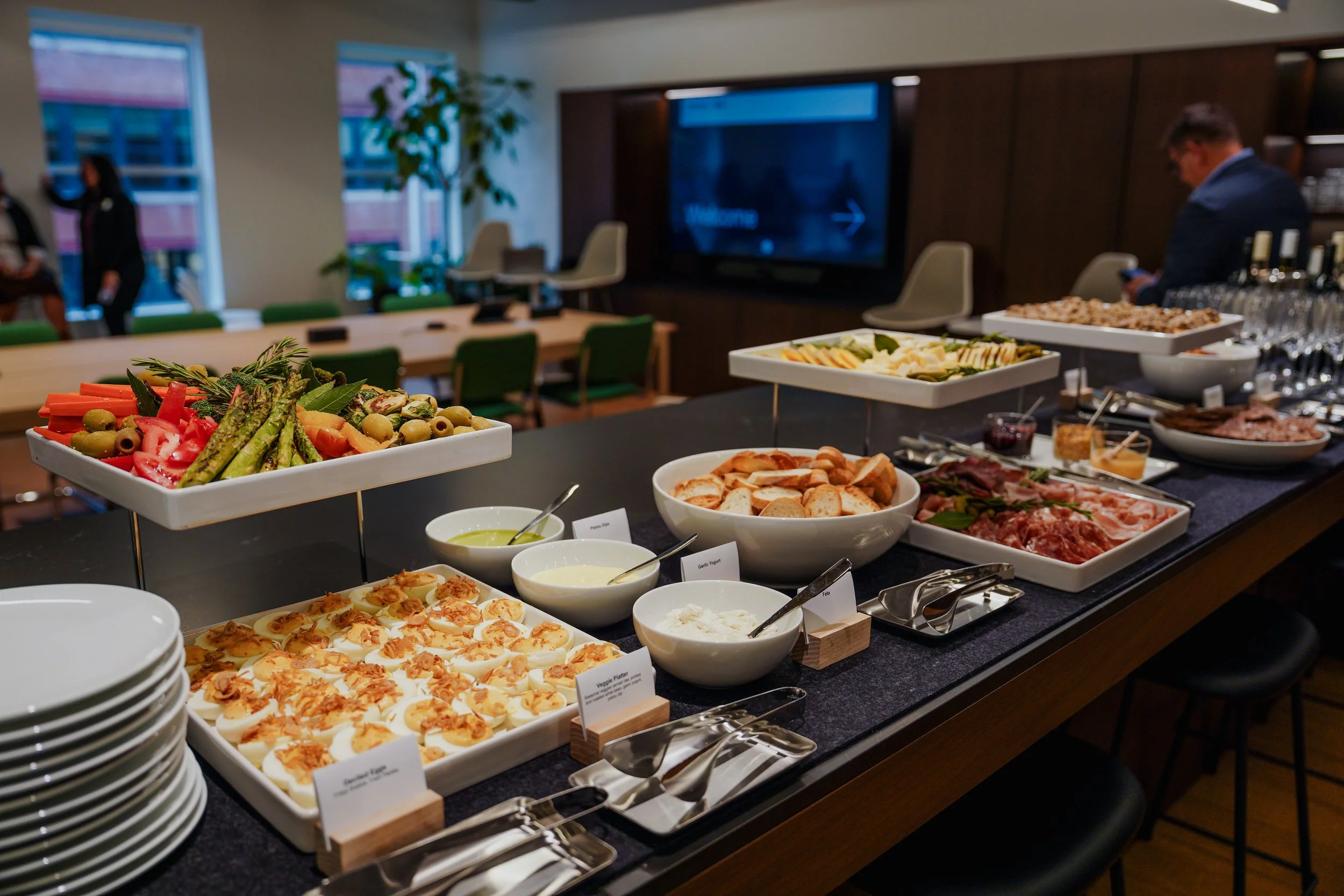 Buffet table with various dishes including deviled eggs, assorted breads, cheeses, cold cuts, salad vegetables, and condiments in a conference room setting with people in the background.