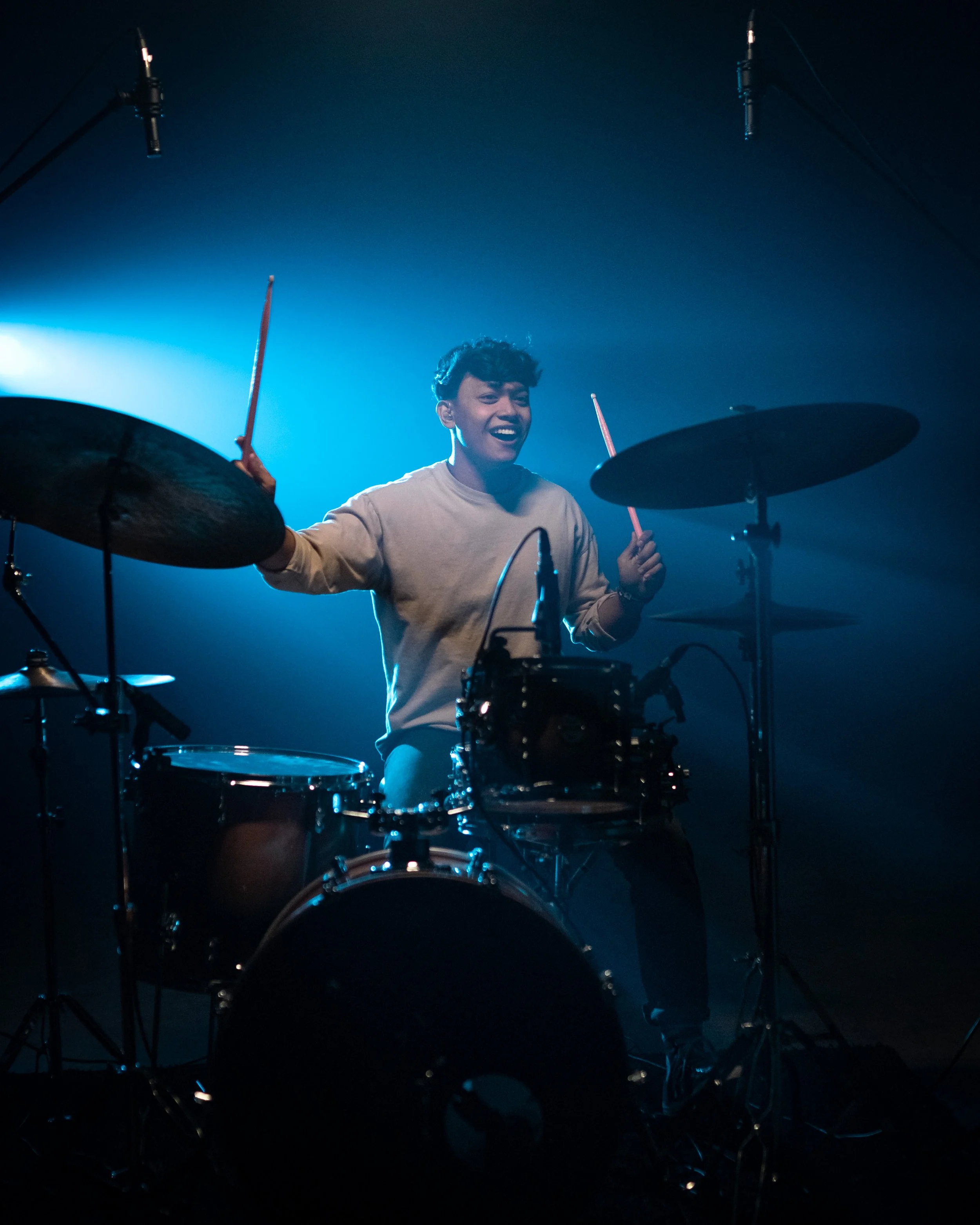 A portrait of a young man playing a drum set on stage, smiling, with blue lighting and microphones overhead.