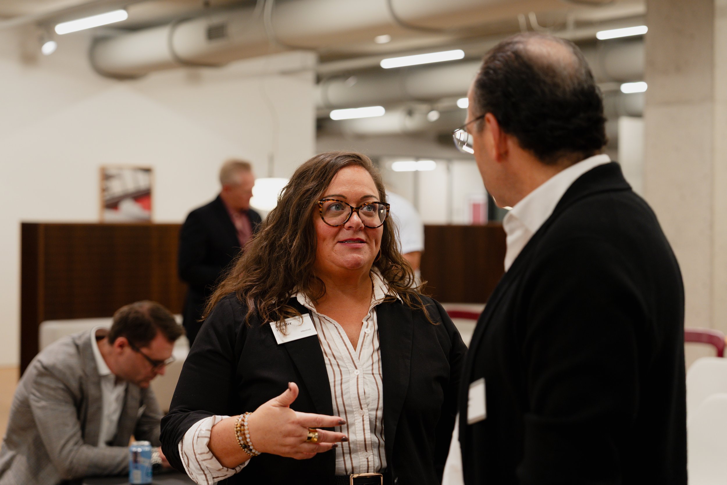 A woman talking to a man in a business setting, with other people in the background.