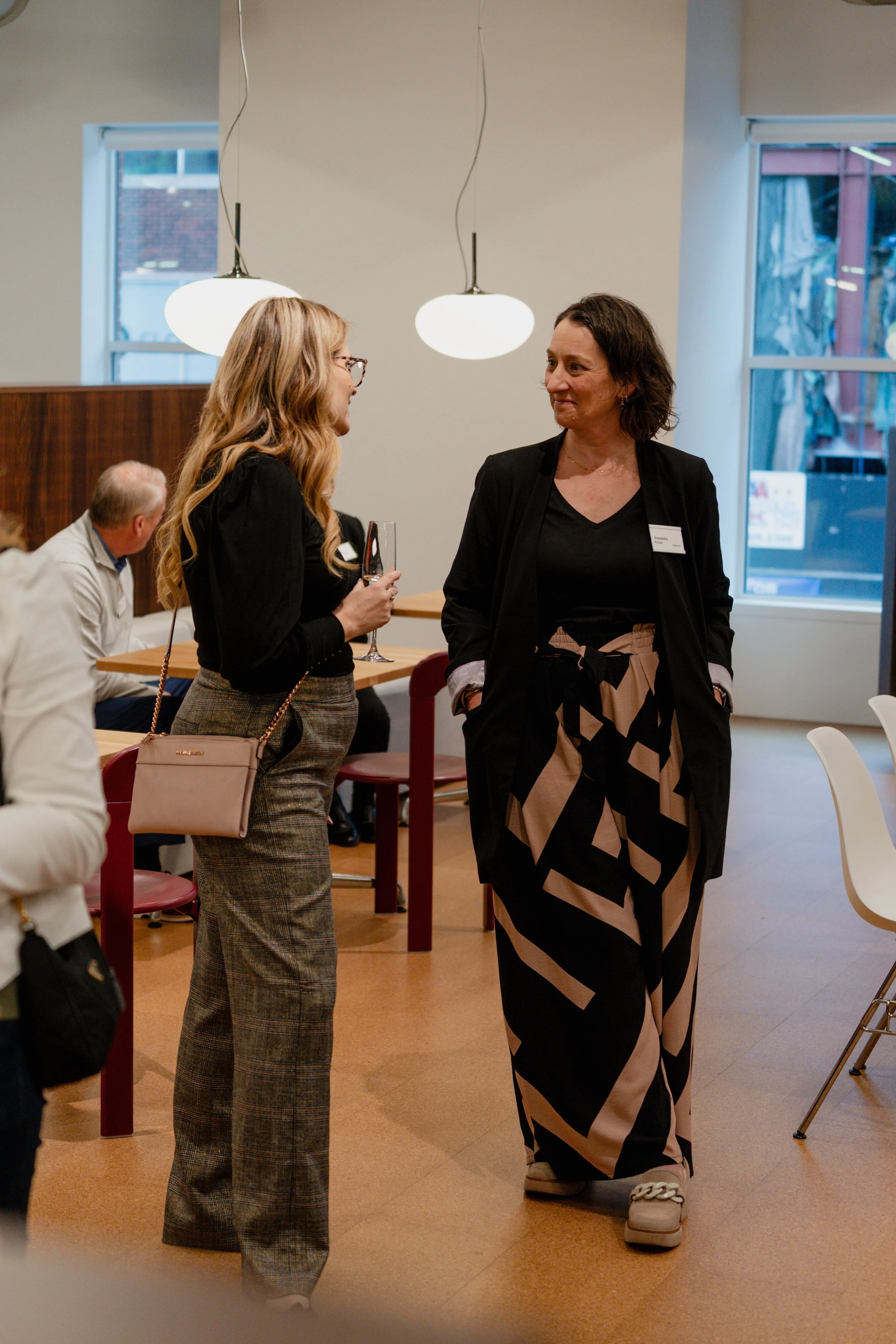 Two women are standing and conversing indoors at a social event, with windows and hanging lights in the background.