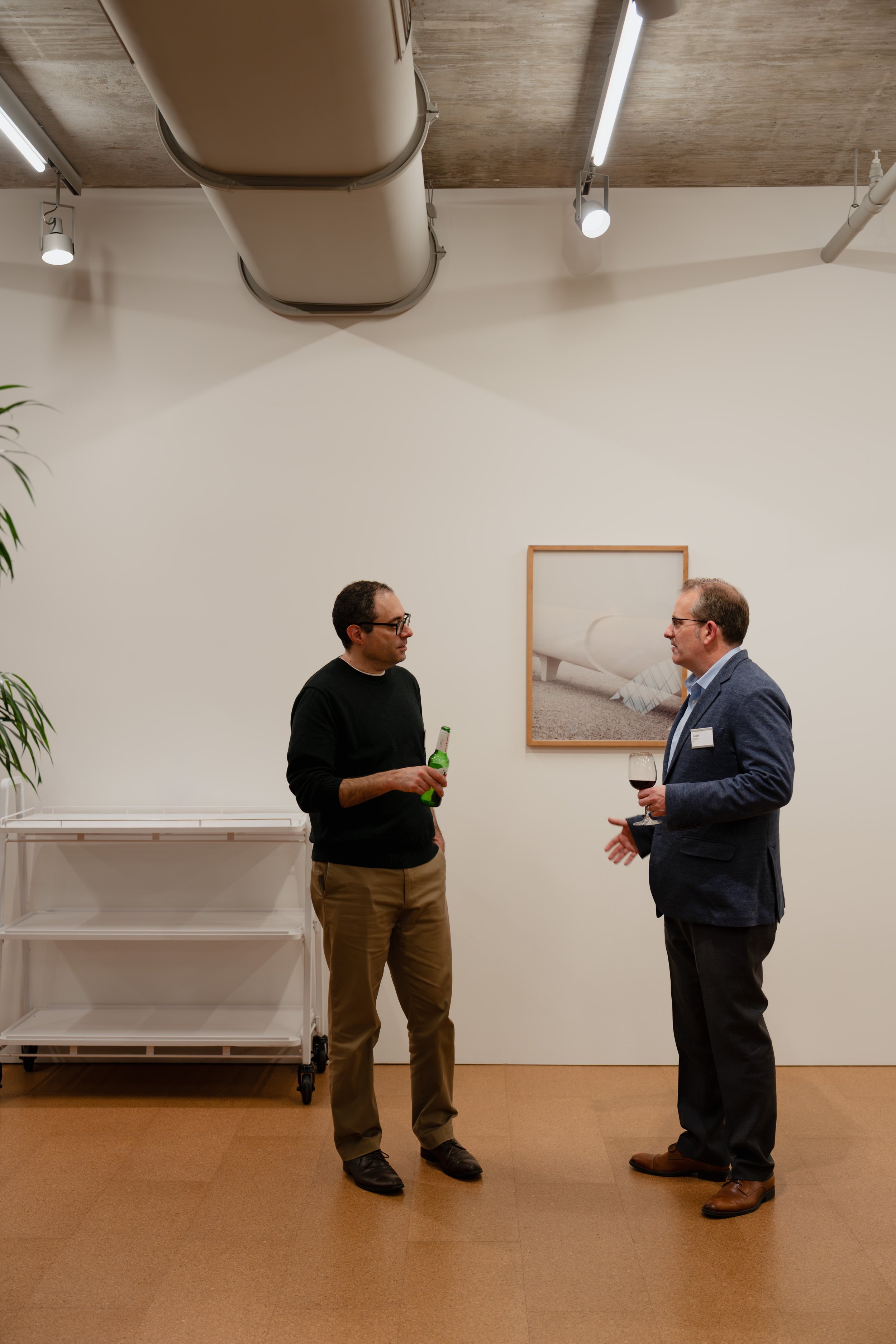 Two men are talking at an art gallery or reception, one holding a beer, the other holding a glass of red wine.
