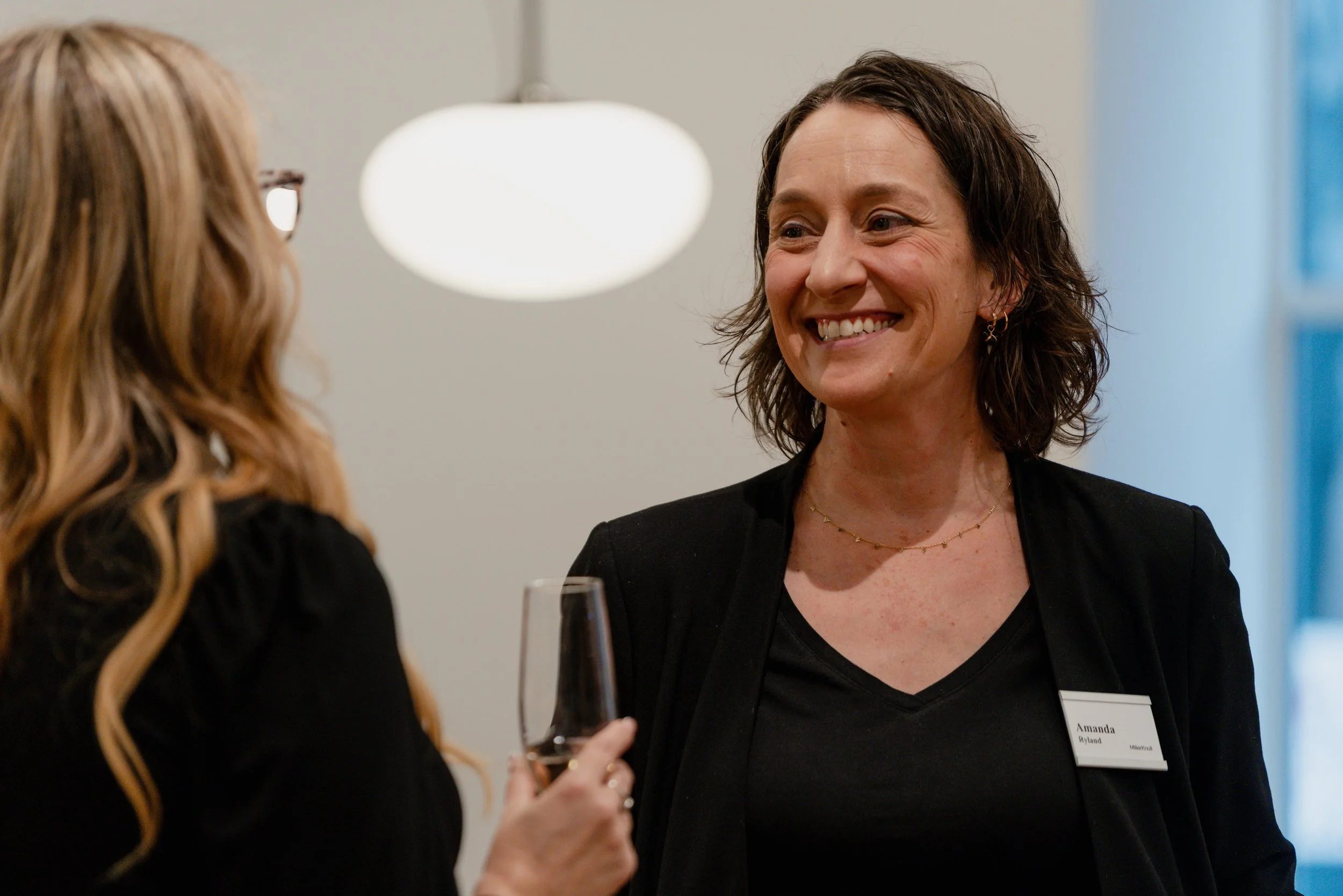 Two women talking at a social event, one holding a glass of wine, smiling, wearing a black blazer and a name tag, in an indoor setting with soft lighting.