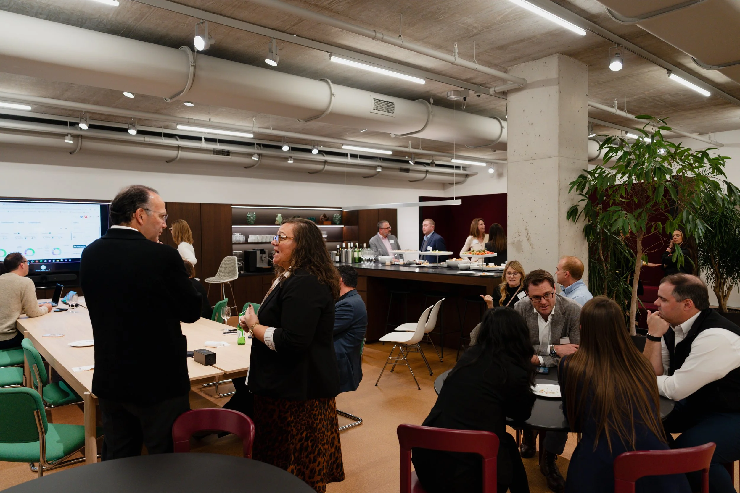 A corporate event or meeting taking place in a modern office space, with people engaging in conversations, a speaker at a black table with food and drinks, and a large screen displaying data in the background.