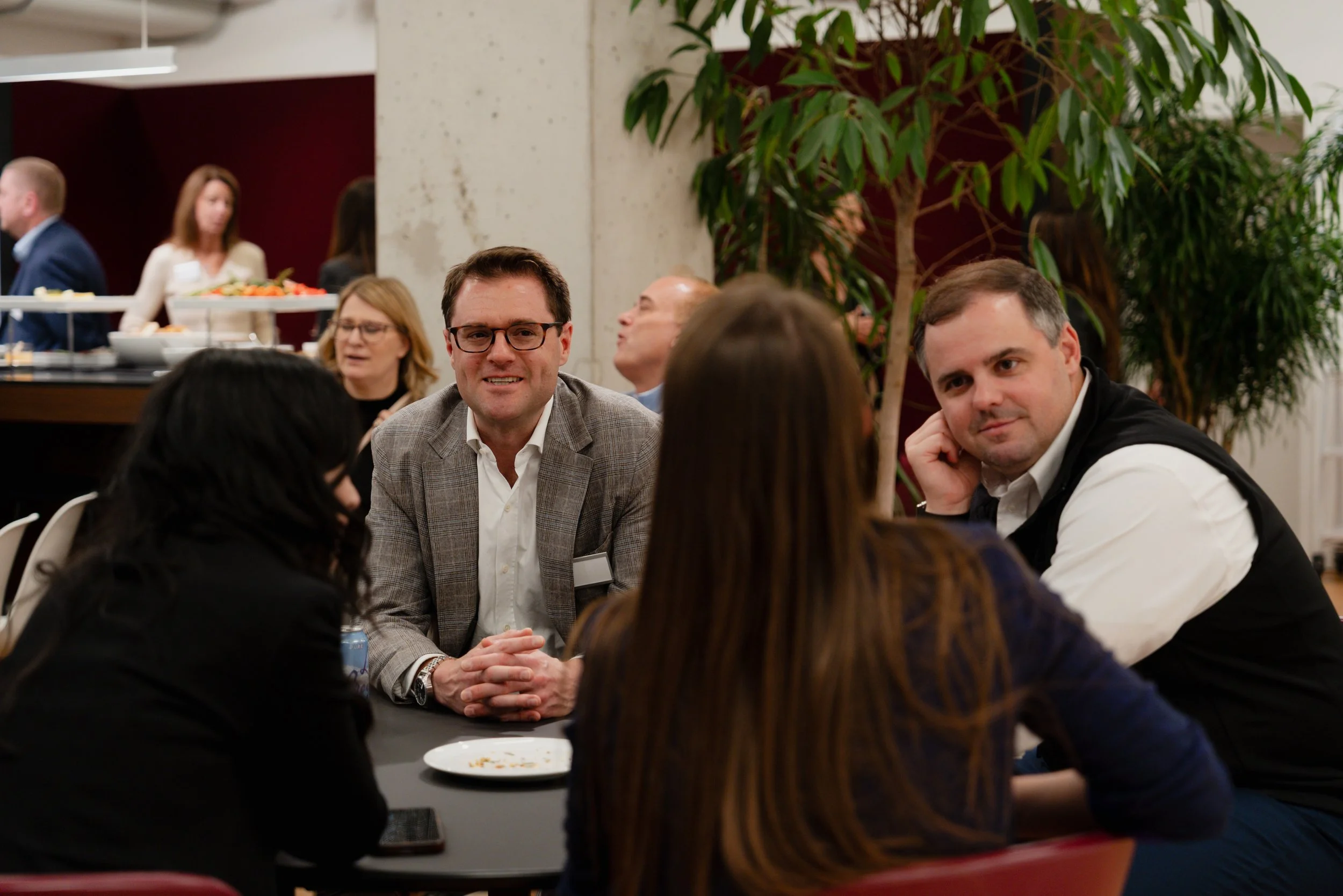 People sitting and talking at a table during a social event or conference, with some people in the background and a large potted plant nearby.