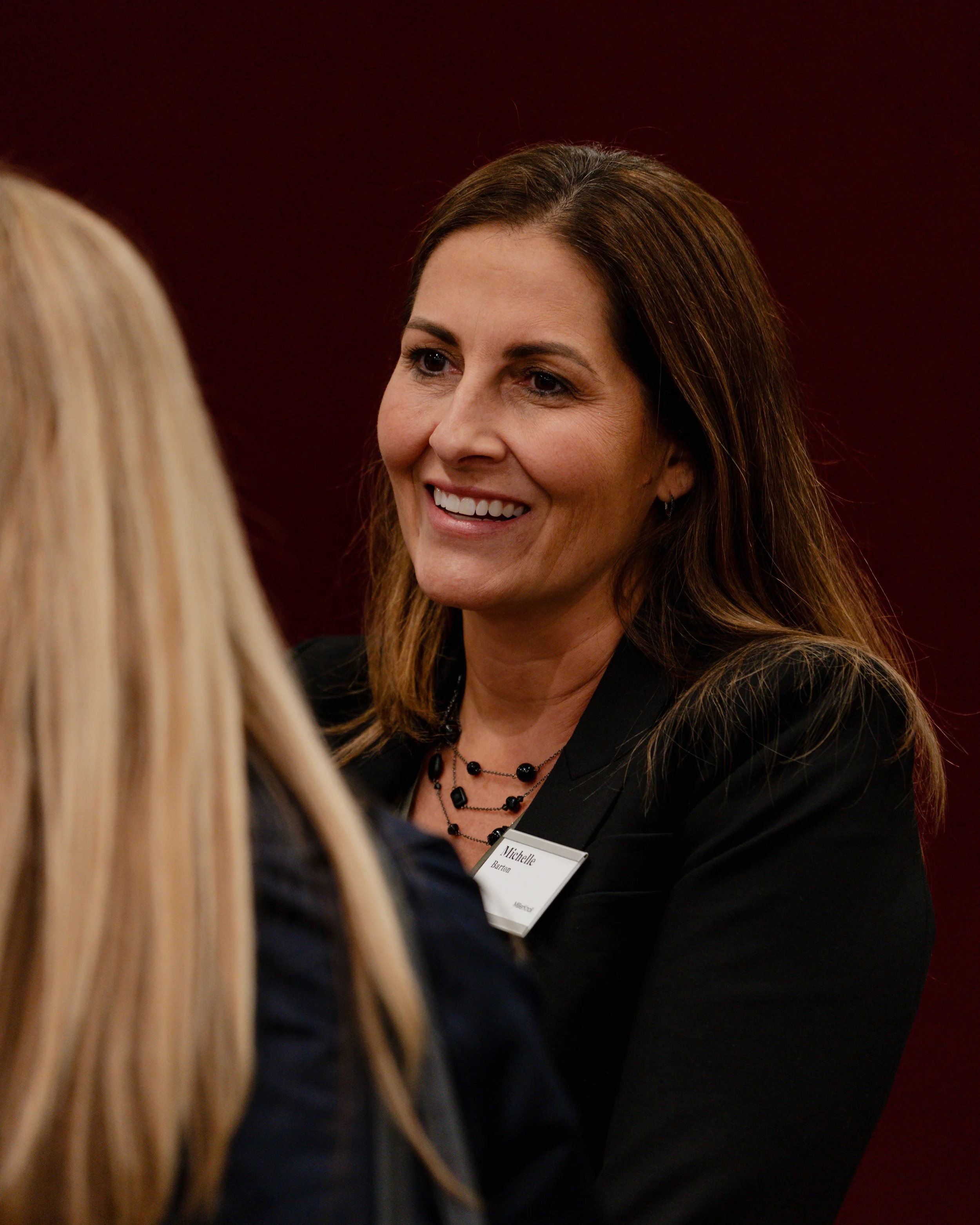 A woman with brown hair smiling and talking to another woman with blonde hair in a professional setting.