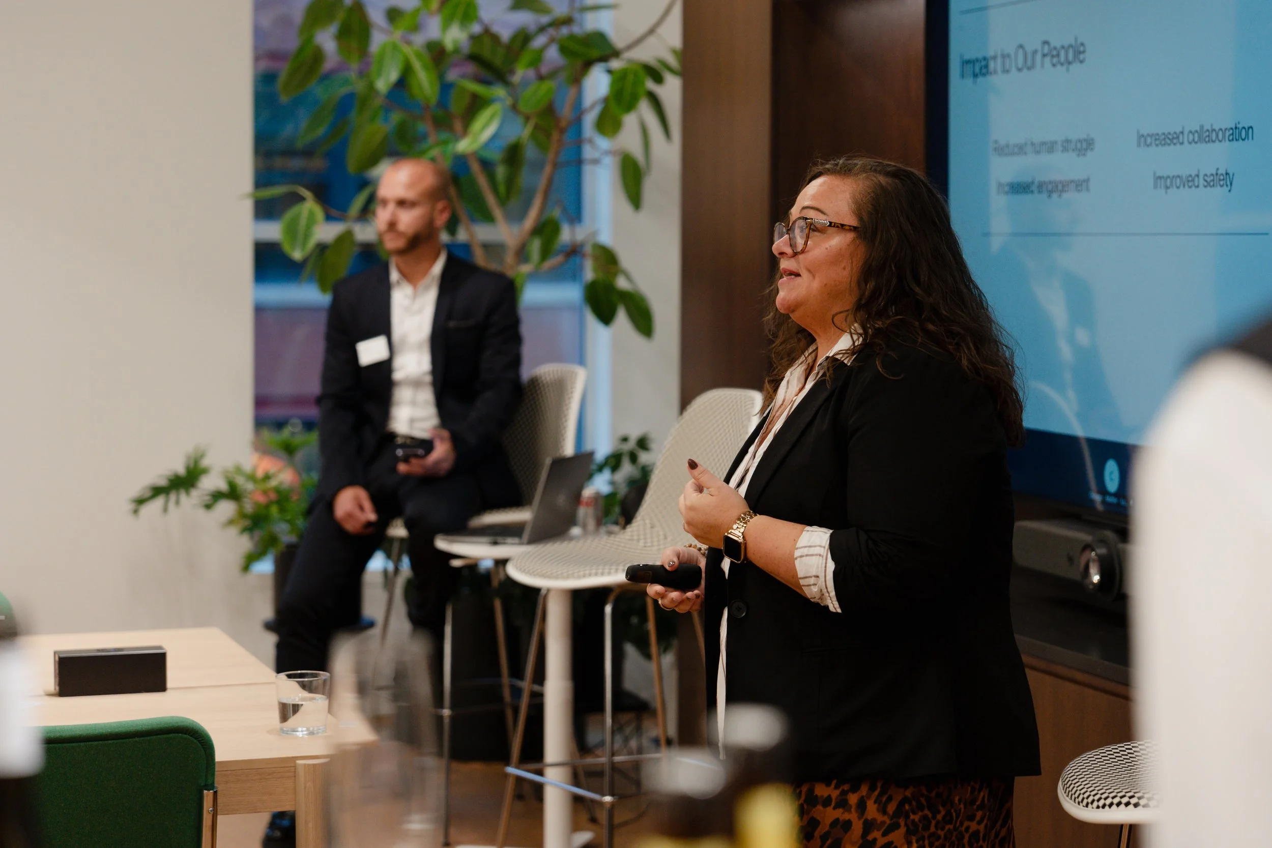 A woman with glasses, a black blazer, and leopard print pants giving a presentation in a conference room, with a large screen behind her displaying a slide titled 'Inspire in Our People.'