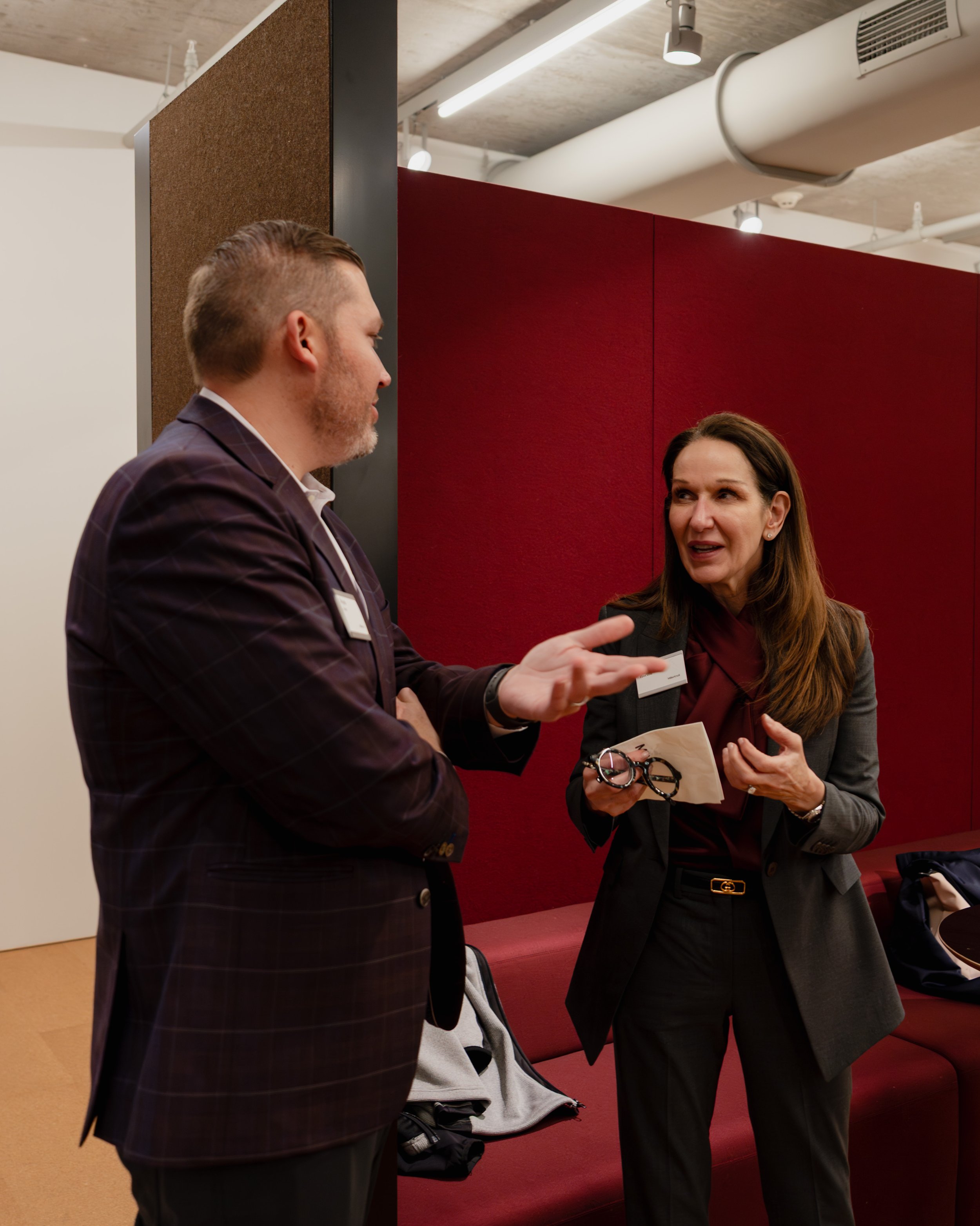 Two business professionals, a man and a woman, engaged in conversation in an office or conference setting. The man is gesturing with his hand while the woman appears to be listening and holding a pair of glasses and a small paper.