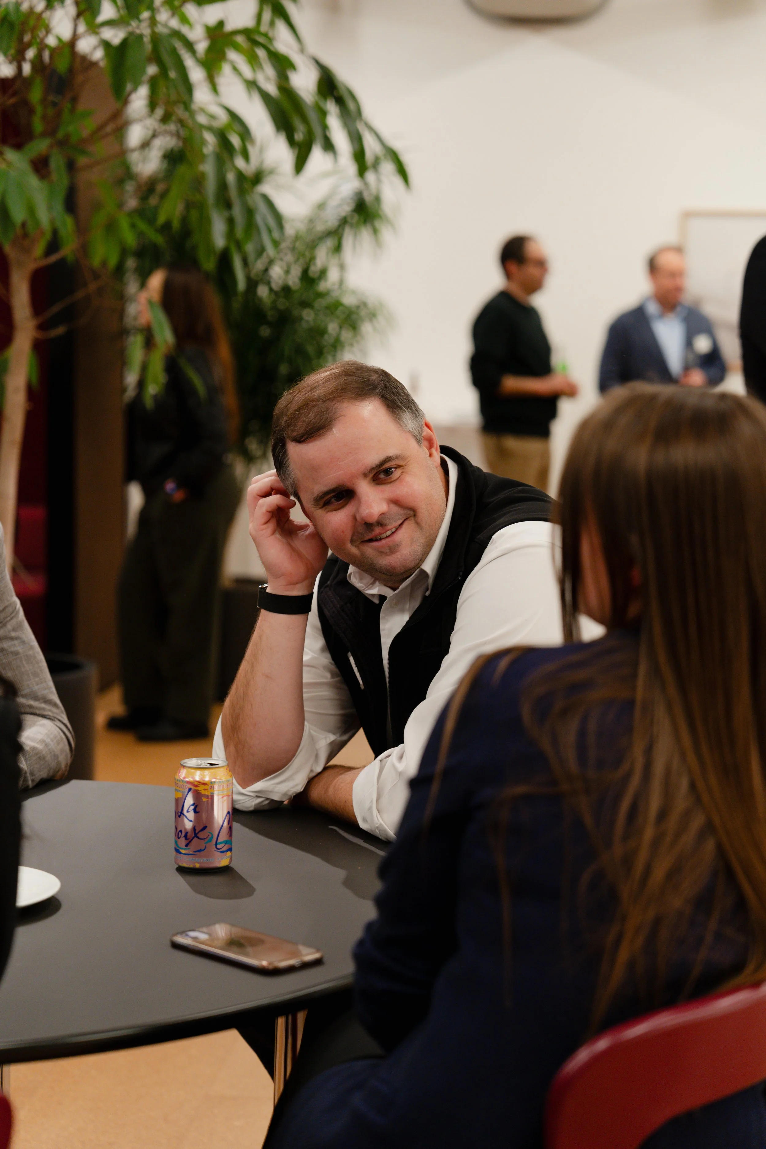 A man and woman sitting at a table in a social event or networking gathering, with the man smiling and engaging in conversation. There are other people standing and chatting in the background, along with a large potted plant and a white wall.