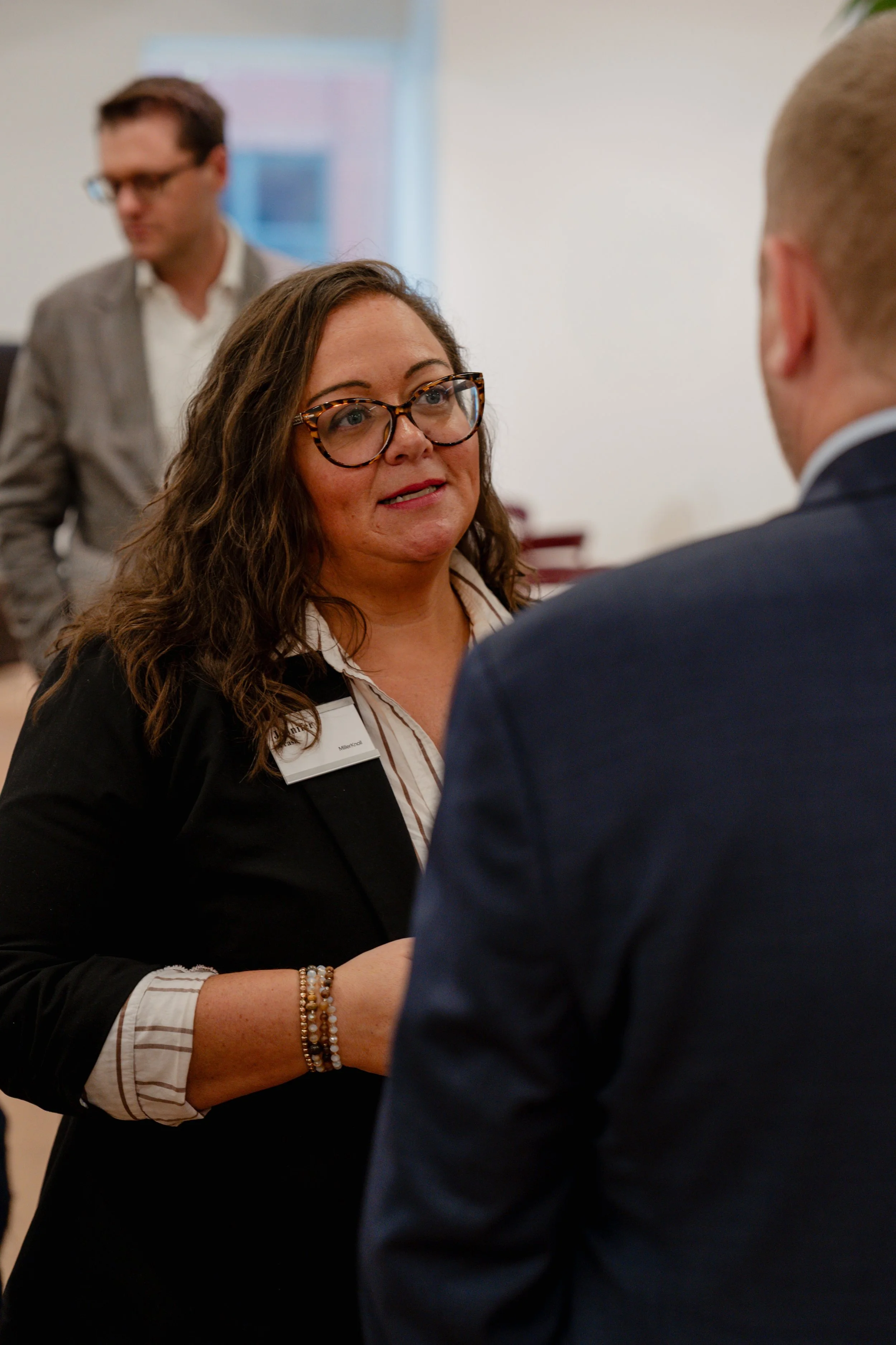 A woman with glasses and a white blouse talking to a man in a dark suit, with another man blurred in the background.
