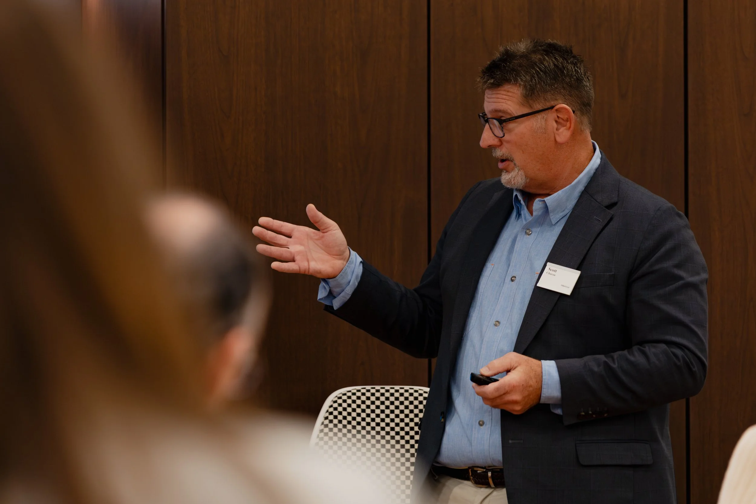A man with glasses and a beard in a dark blazer and light blue shirt is speaking, gesturing with his right hand, at a professional event or presentation in a room with wooden walls.