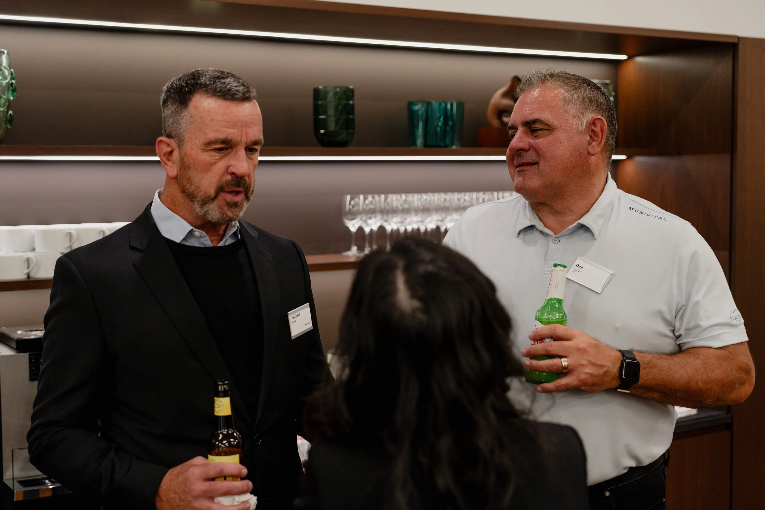 Two men and a woman at a social gathering. The man on the left has gray hair, a beard, and is wearing a black suit, holding a beer bottle. The man on the right has short gray hair, wearing a white shirt, and holding a green bottle. The woman, with da