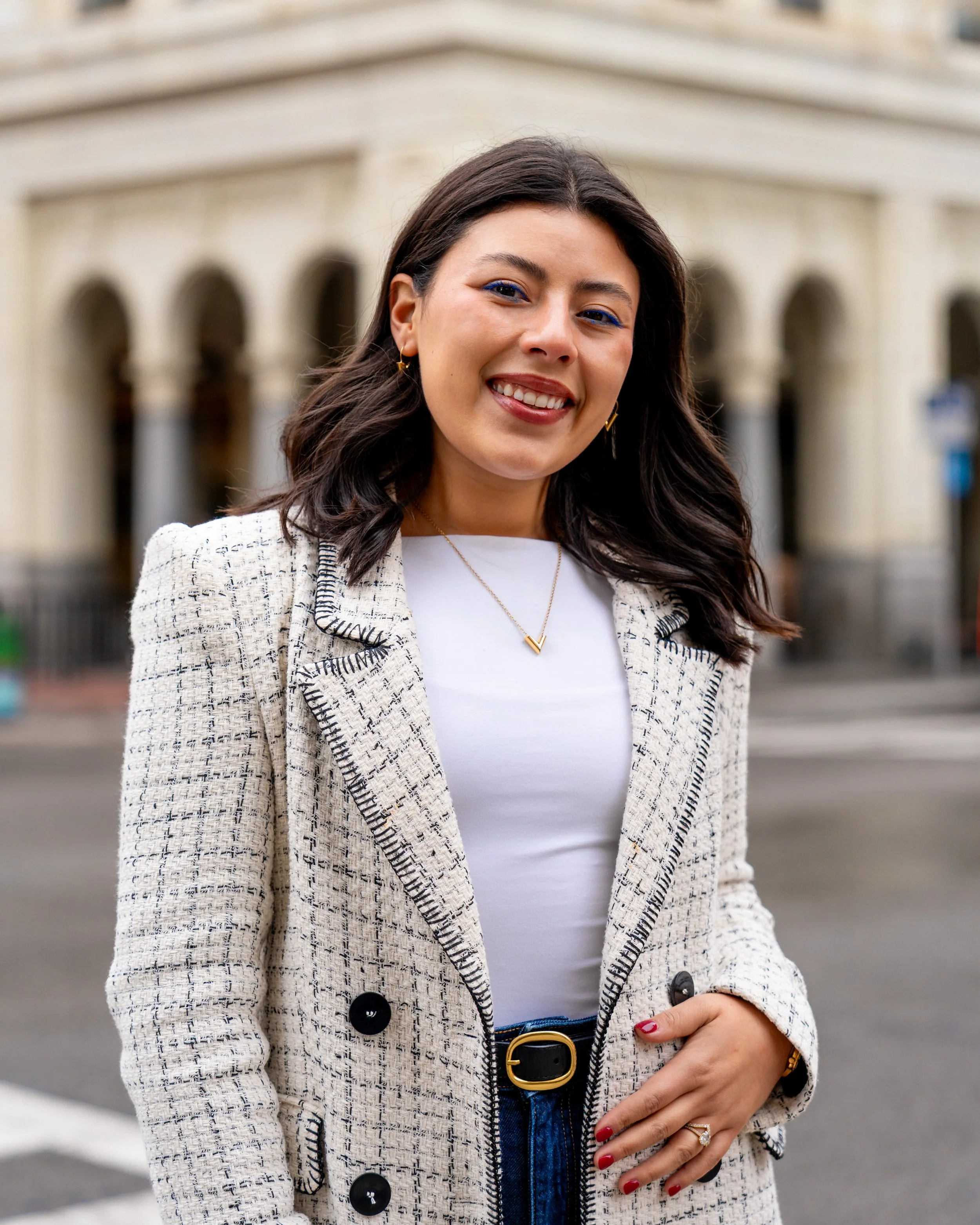 A smiling woman with dark brown hair styled in loose waves, wearing a white t-shirt, a beige plaid blazer with black buttons, and blue jeans with a black and gold belt, standing outdoors in front of an ornate building with arches.