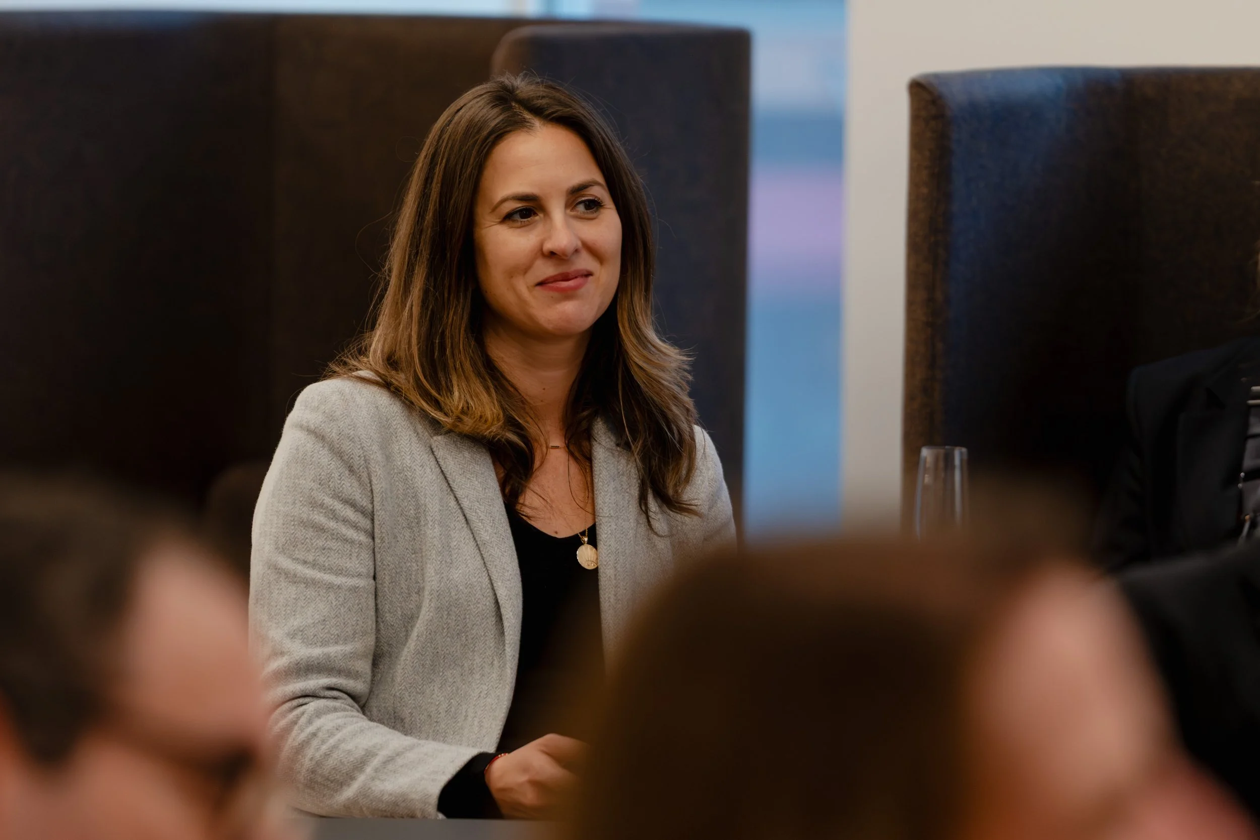 Woman with brown hair, wearing a light gray blazer and black top, sitting at a table during a meeting or event.