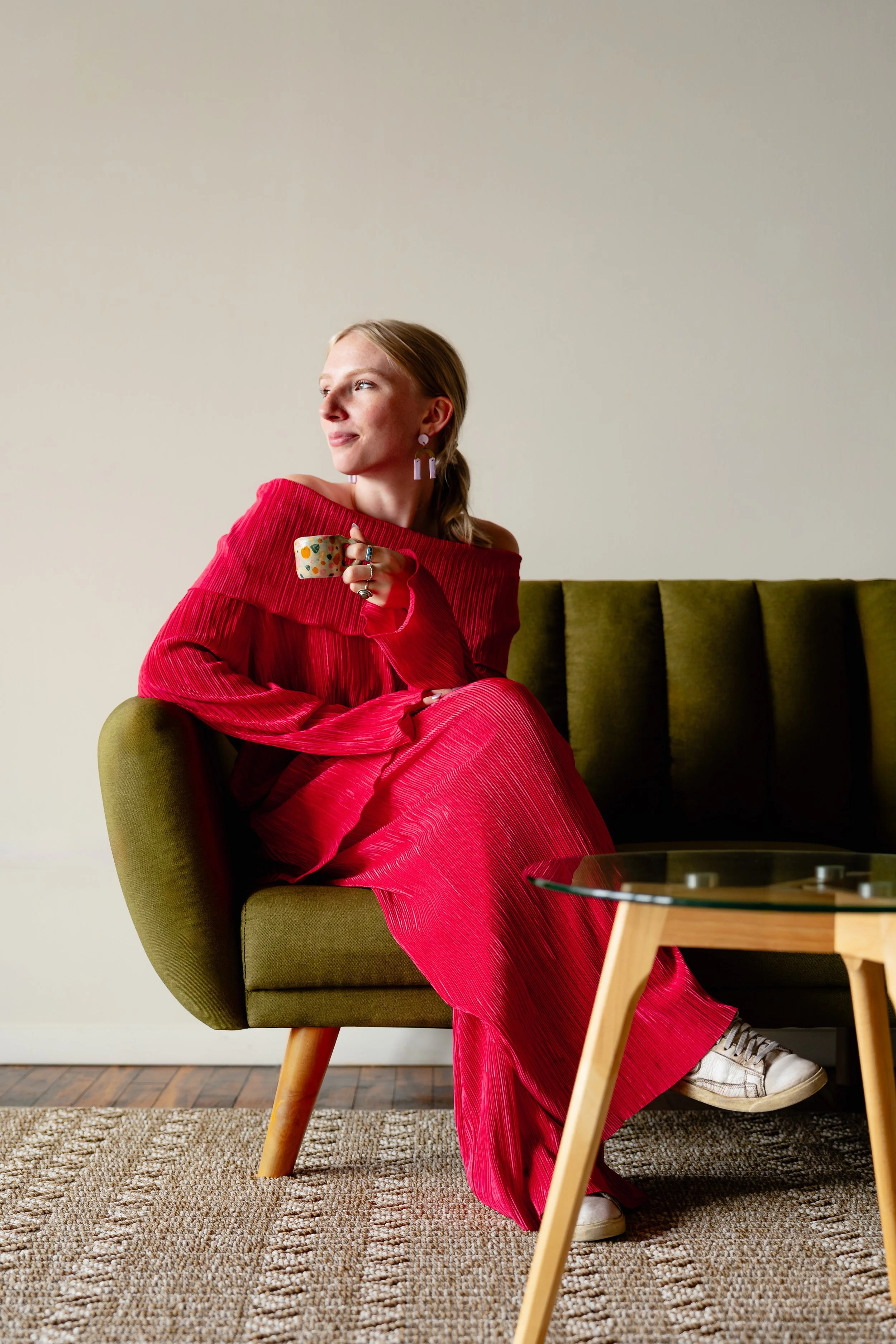 A portrait of a woman in a red pleated dress sitting on a green sofa, holding a mug, with braided hair, wearing statement earrings and white sneakers.