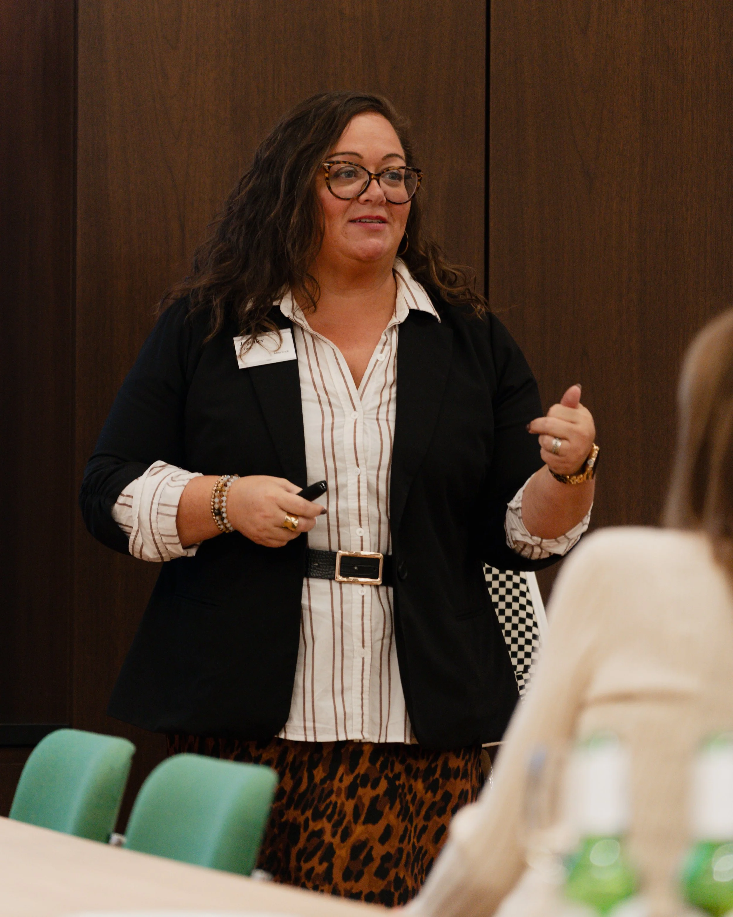 Woman with glasses and curly hair speaking at a meeting or presentation, holding a remote control, wearing a black blazer, striped shirt, and animal print skirt, in a conference room.