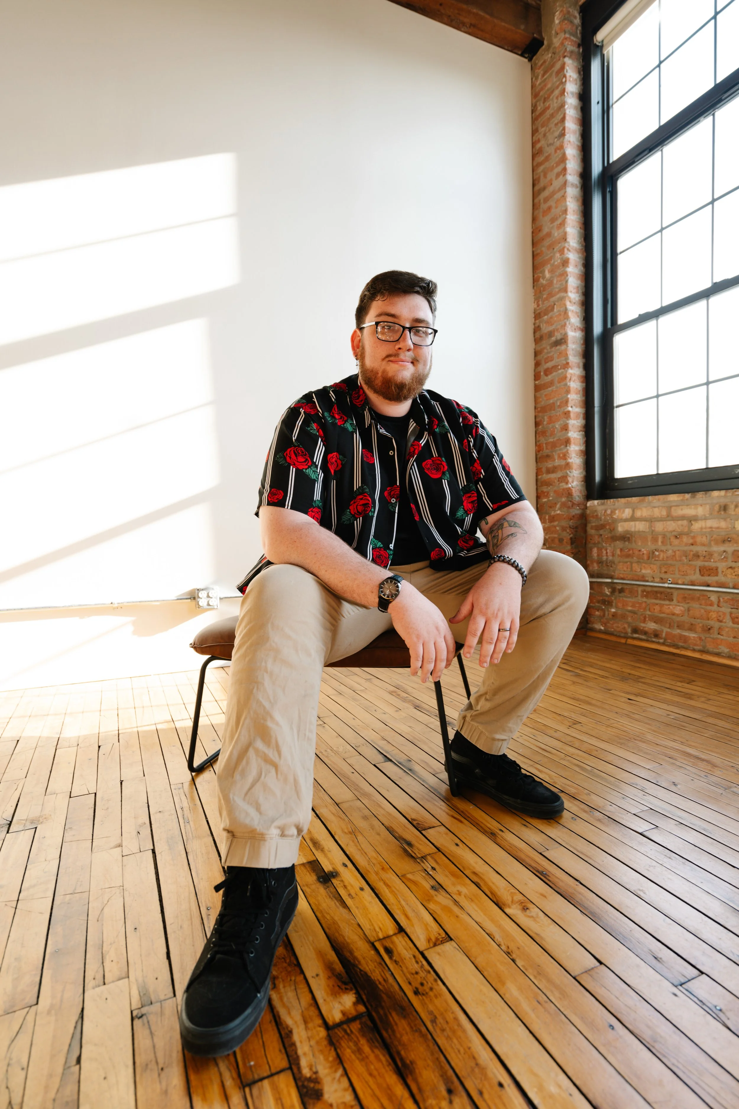 A portrait of a man with glasses, a beard, wearing a black shirt with red roses, khaki pants, and black sneakers, sitting on a chair in a room with wooden floors, brick walls, and large windows letting in sunlight.
