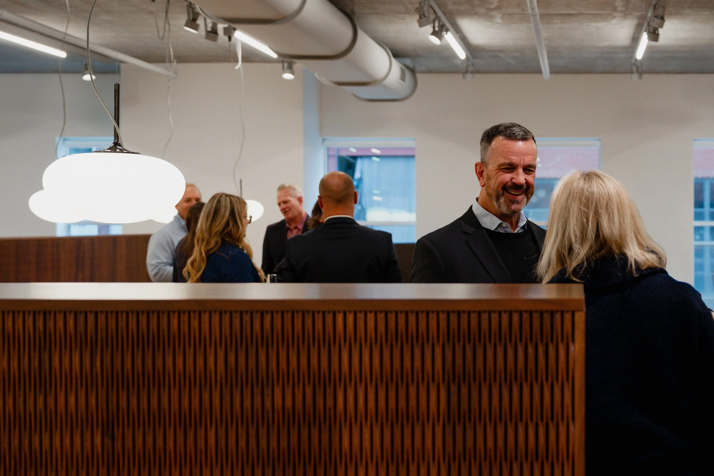 People gathered in a modern office space, engaging in conversation and smiling, with prominent hanging lights, large windows, and a wooden divider.