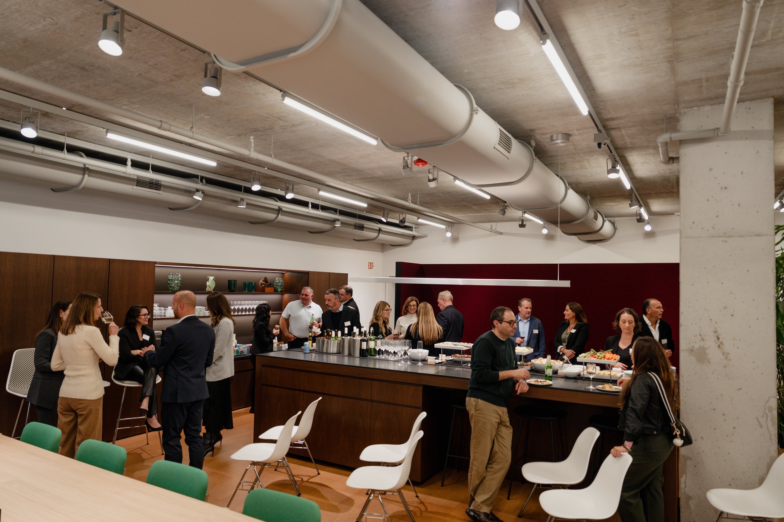 People gathered in a modern, industrial-style room with a concrete ceiling and exposed ductwork, socializing around a large island bar with drinks and snacks, some sitting on white chairs, others standing and talking.
