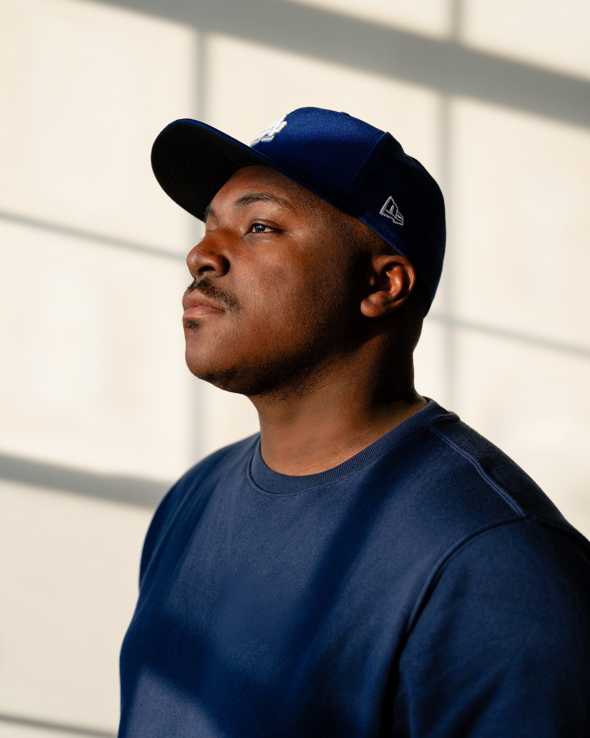 A portrait of a man wearing a blue baseball cap and a blue shirt, looking confidently to the side, with sunlight streaming through window shutters in the background.