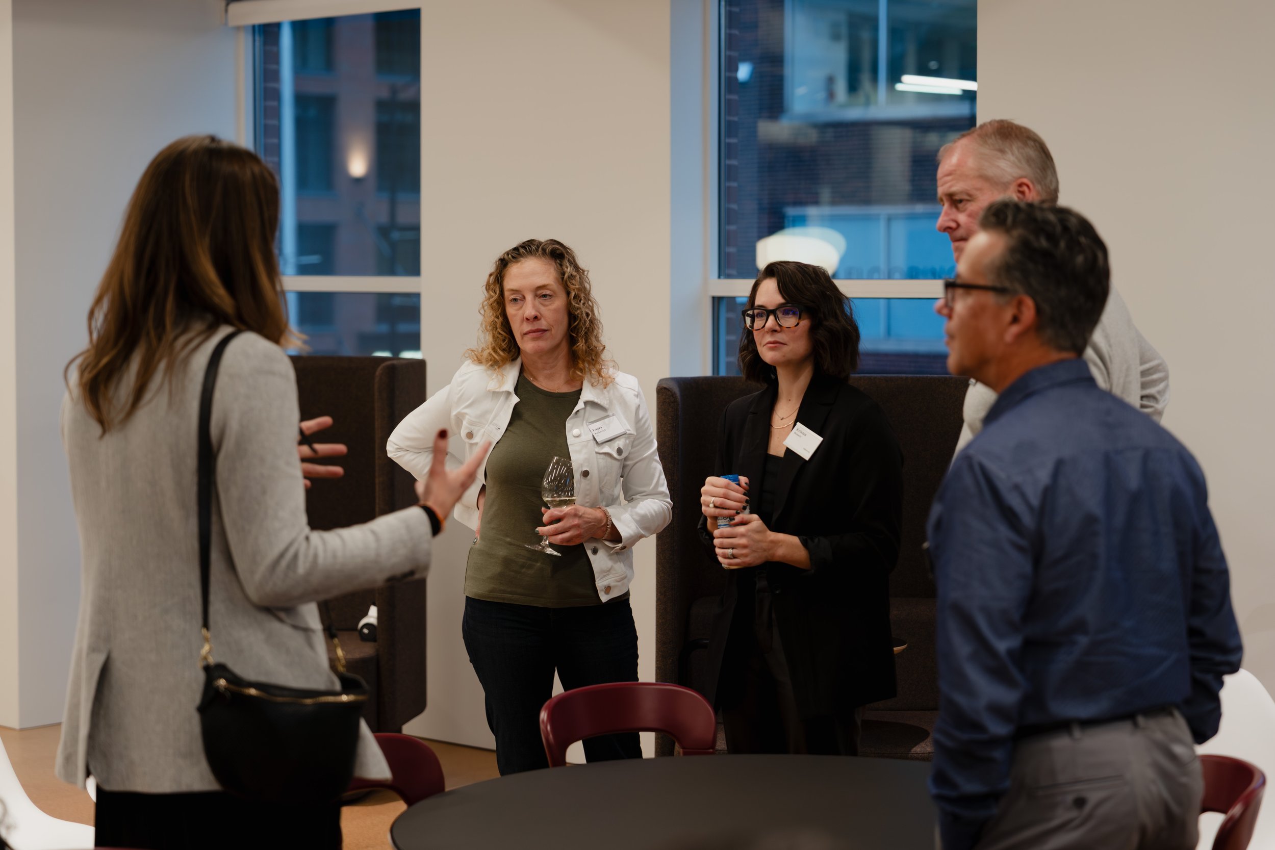Group of five people in business attire having a conversation in a modern office with windows showing city buildings.