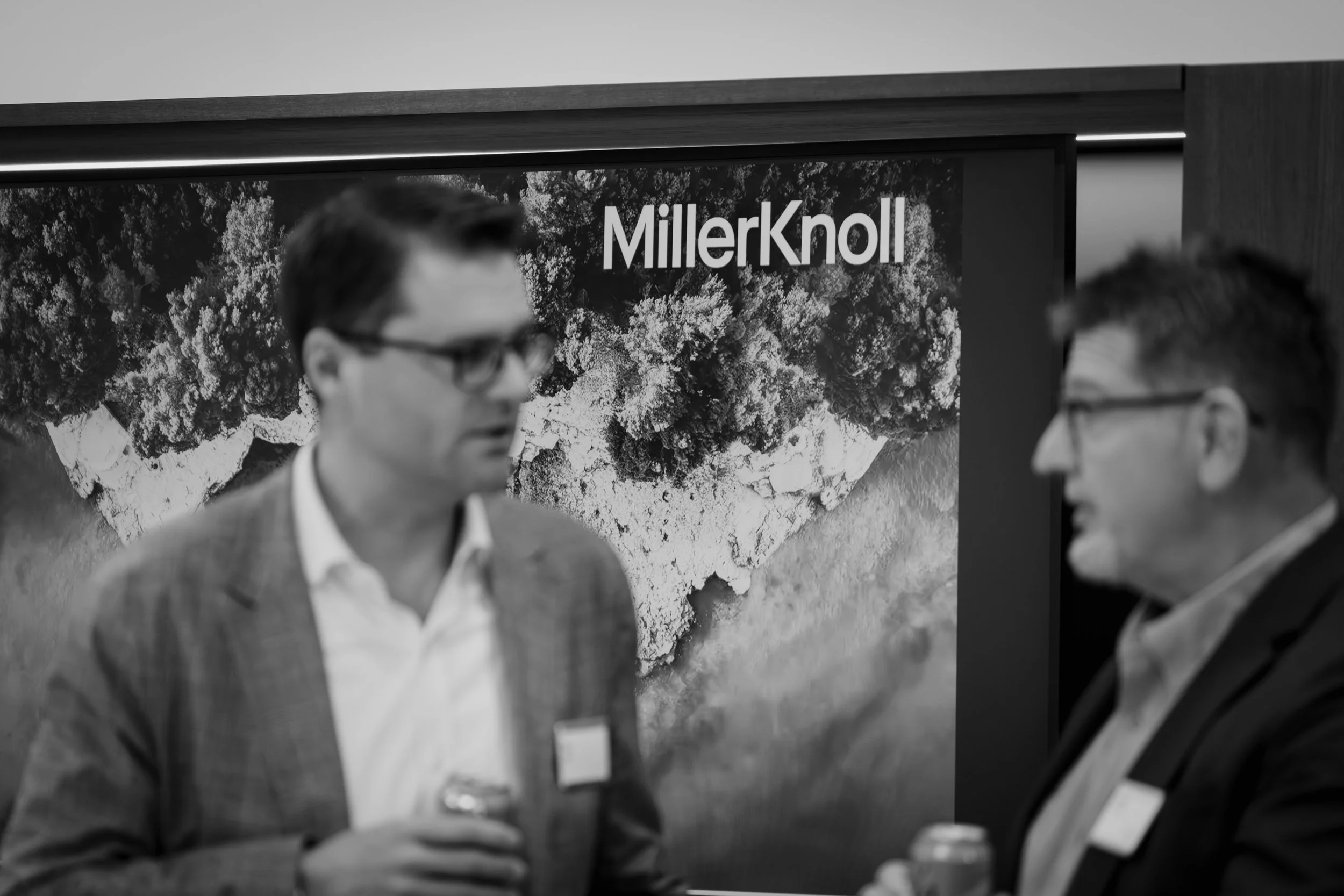 Two men in business attire are talking in front of a MillerKnoll display with an aerial landscape photograph behind them.