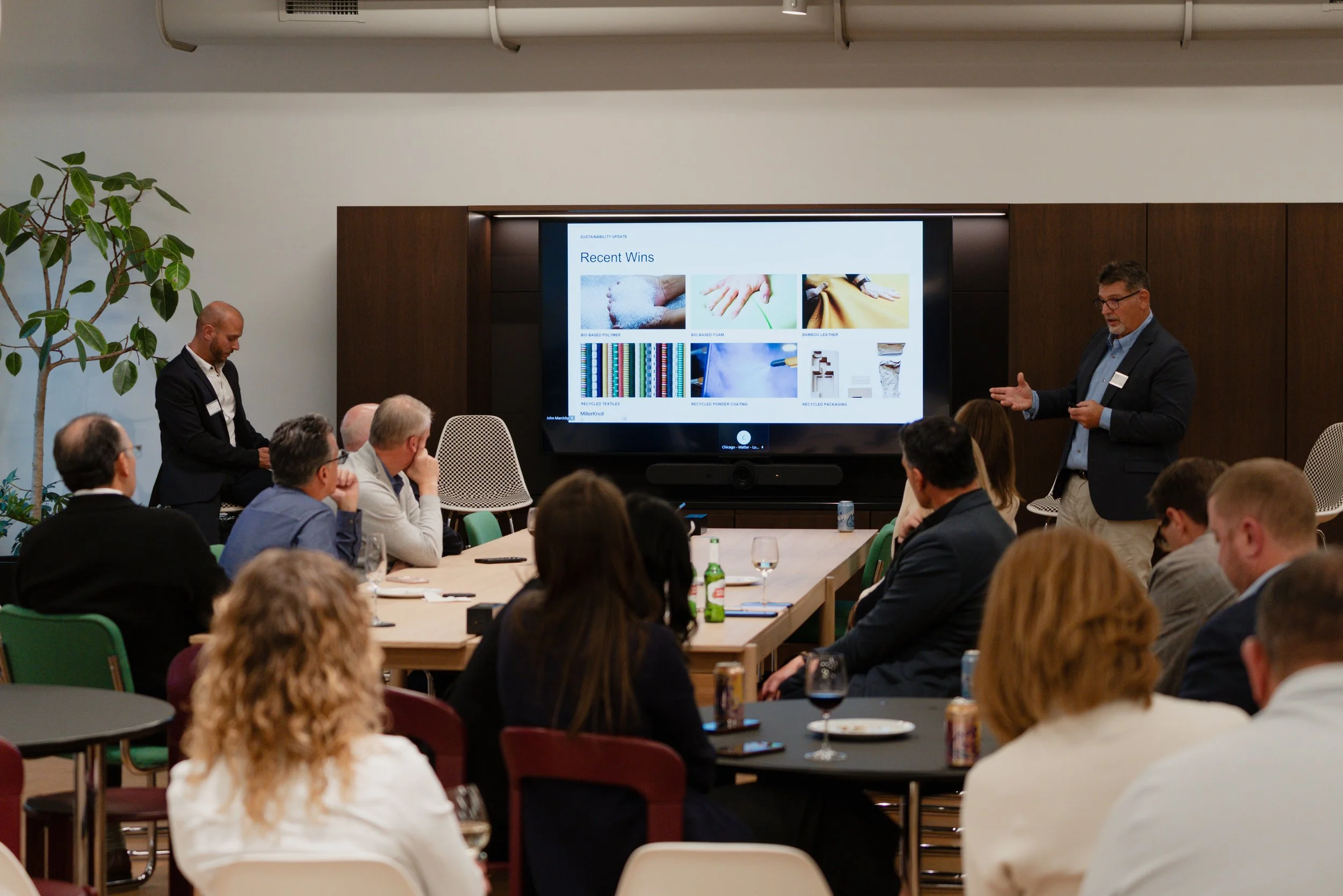 A man giving a presentation in a conference room with a large screen displaying slides titled 'Recent Wins' and images of textiles and recycled materials, while an audience listens.