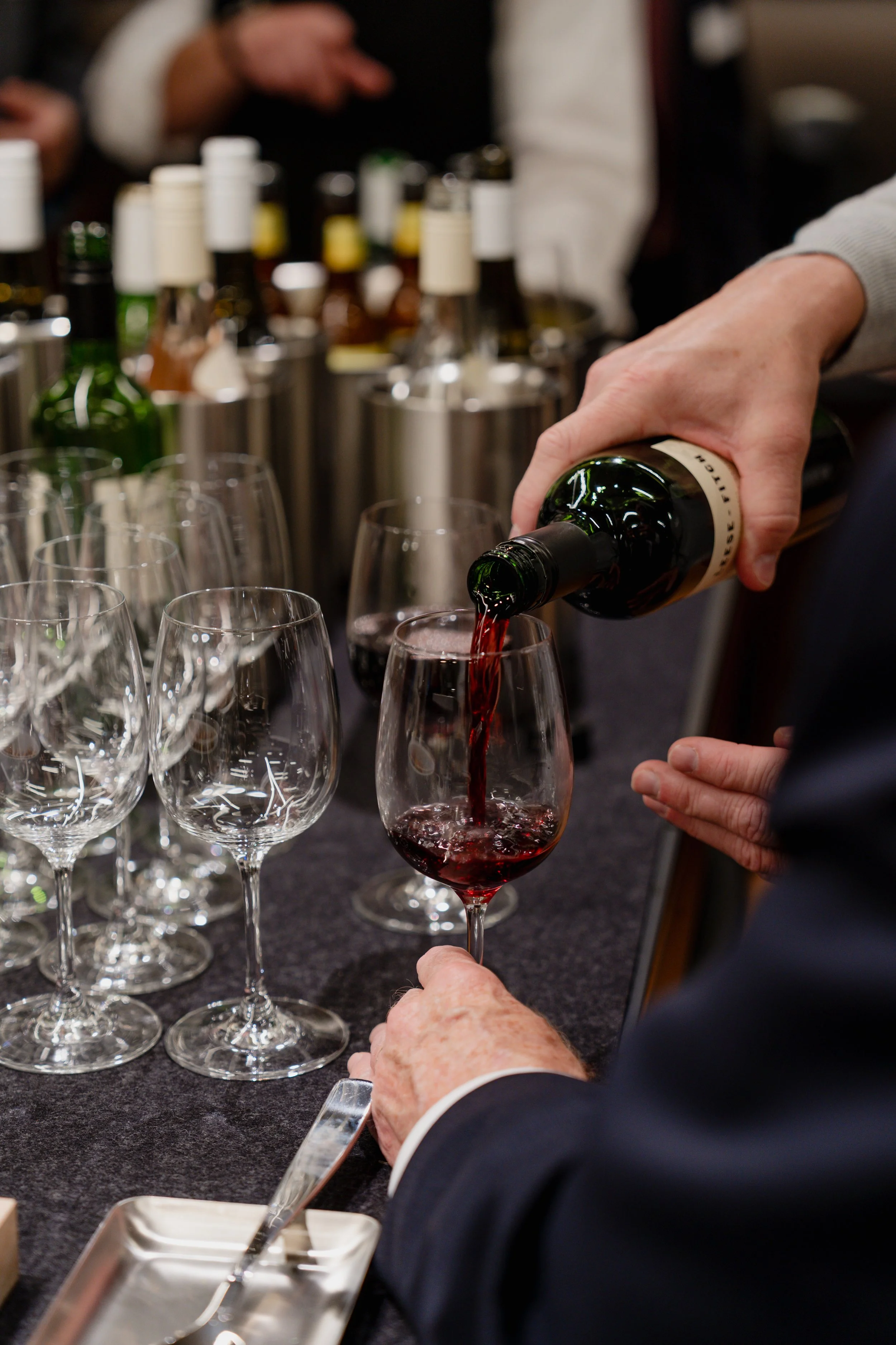 Person pouring red wine into a glass at a tasting event, with several empty wine glasses and bottles in the background.