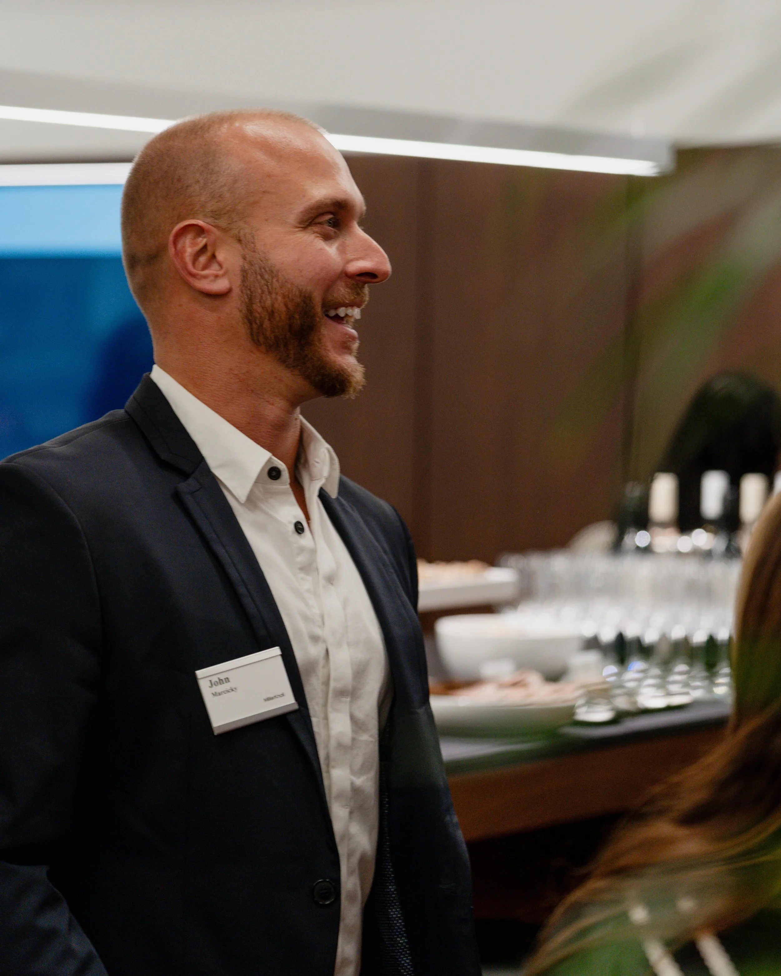 Profile of a smiling man wearing a dark suit jacket and a white shirt, with a name tag reading 'John'. Background features a buffet station with glassware and bowls.